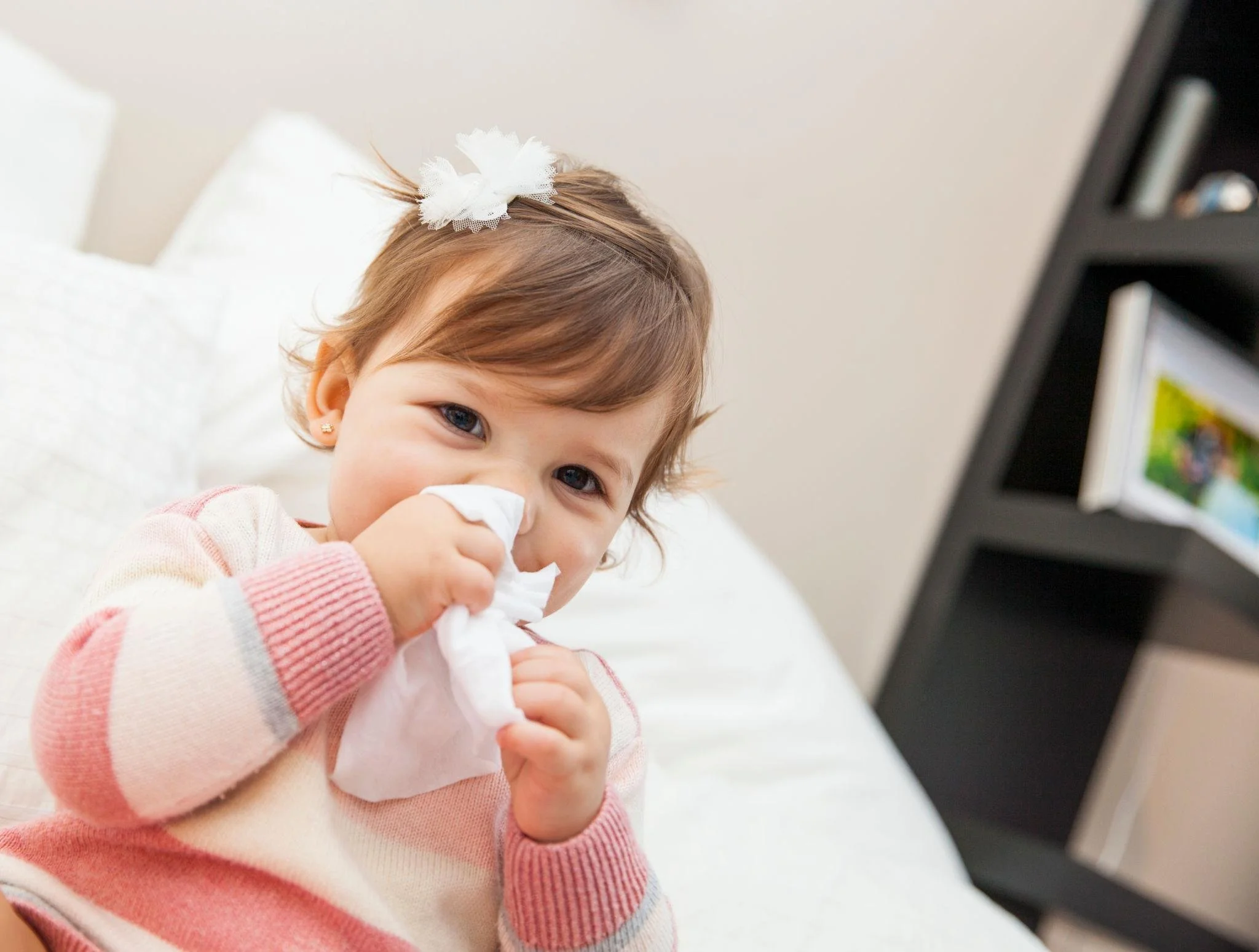 Young girl with brown hair and blue eyes, wearing pink and white striped sweater, holding tissue near nose, with a white bow in her hair, sitting on a white bed near a black shelf with books.