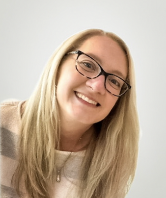 A smiling woman with long blonde hair, glasses, and a light-colored top, standing against a plain light background.