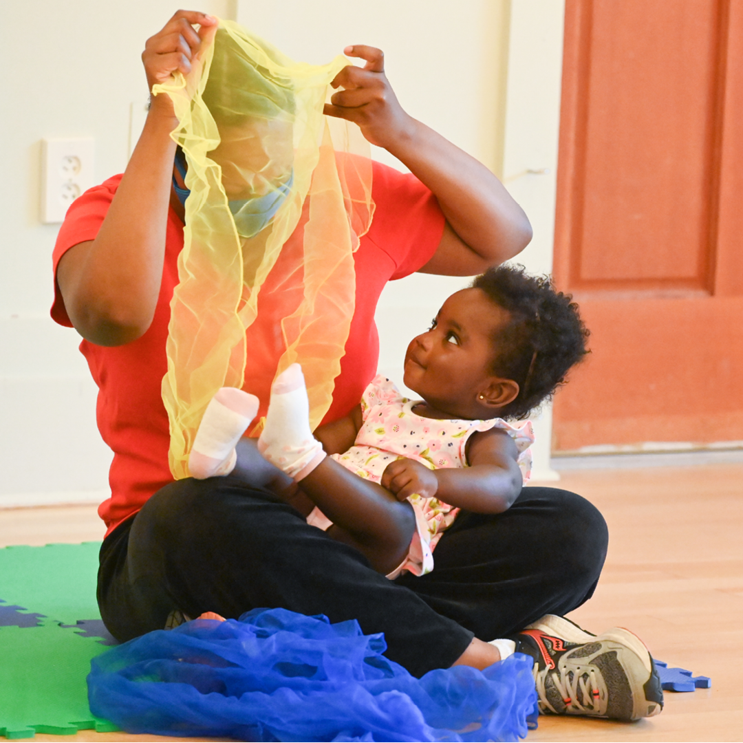 A woman sitting on the floor holding a young girl. The woman is partially obscured by a yellow, semi-transparent fabric, which she is holding above her head. The girl is looking up at the woman with admiration, and they are sitting on a colorful foam mat.