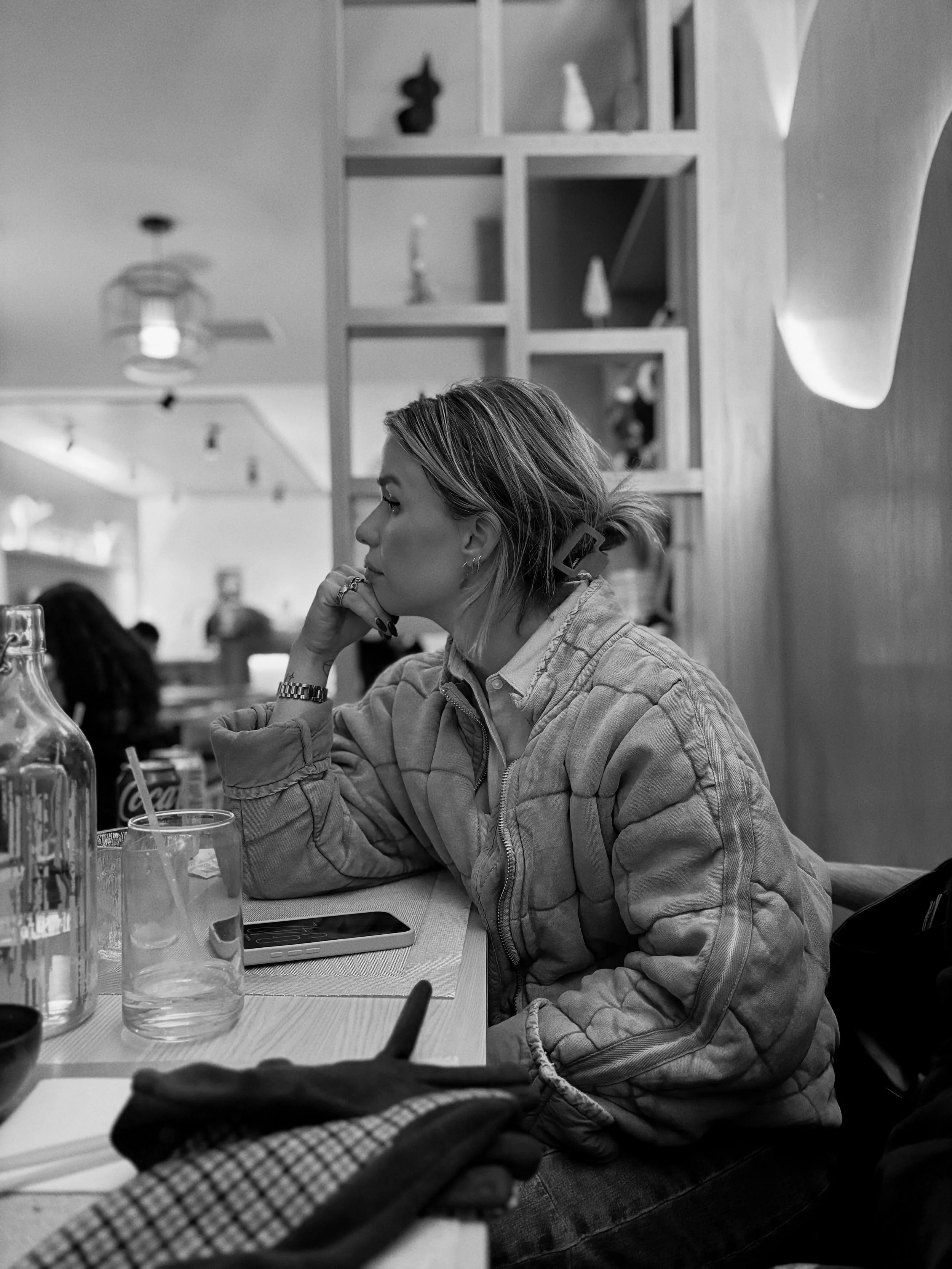 A woman in a plaid shirt sits at a restaurant table, resting her chin on her hand, with a serious expression. The table has water glasses, a phone, and bottles, and a bookshelf with decorative items is behind her. The image is in black and white.