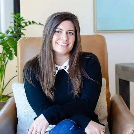 A young woman with long brown hair sitting in a modern office setting, smiling, wearing a black top with a white collar, next to a potted plant and a beige cushion.
