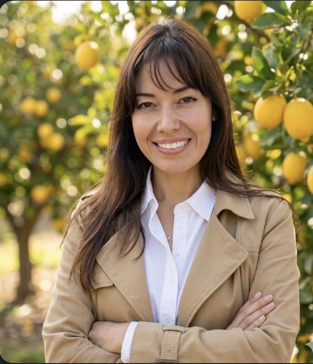 A woman with long brown hair smiling outdoors, standing with arms crossed in front of lemon trees.