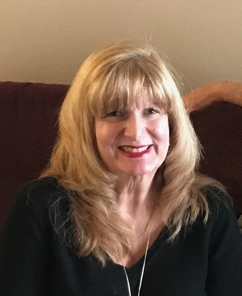 A woman with blonde hair and a black top smiling at the camera, sitting on a dark-colored couch.