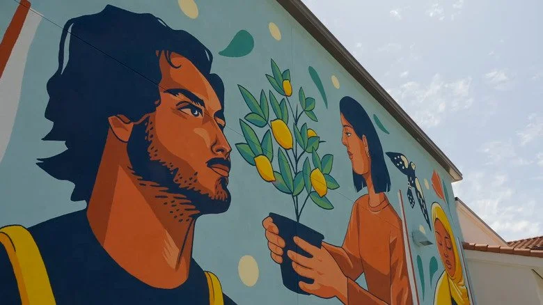Colorful mural of a man with dark hair and beard and a woman with dark hair holding a potted plant with yellow fruit, a butterfly, and a sun. Bright, vibrant artwork.