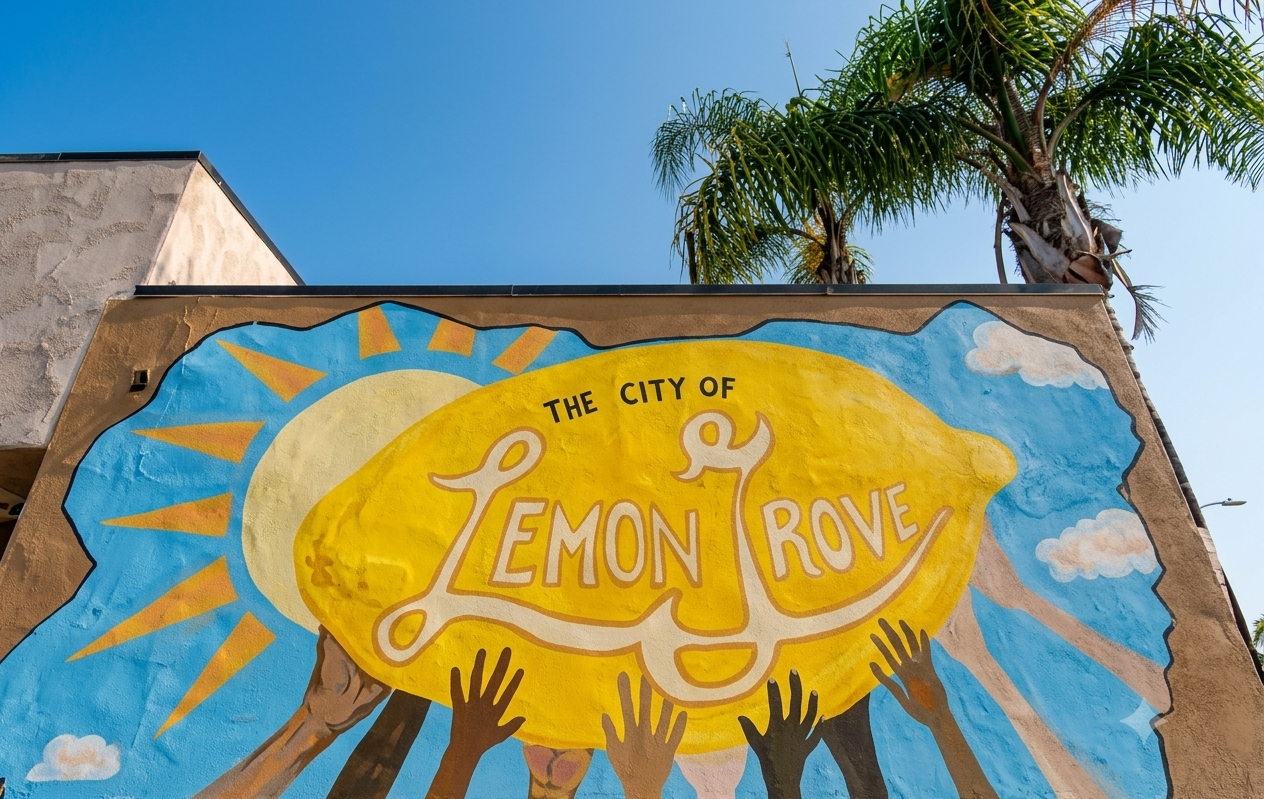 Colorful mural with a yellow lemon in the center, surrounded by raised hands, a sun with rays, and clouds, featuring text that reads 'The City of Lemon Grove'.