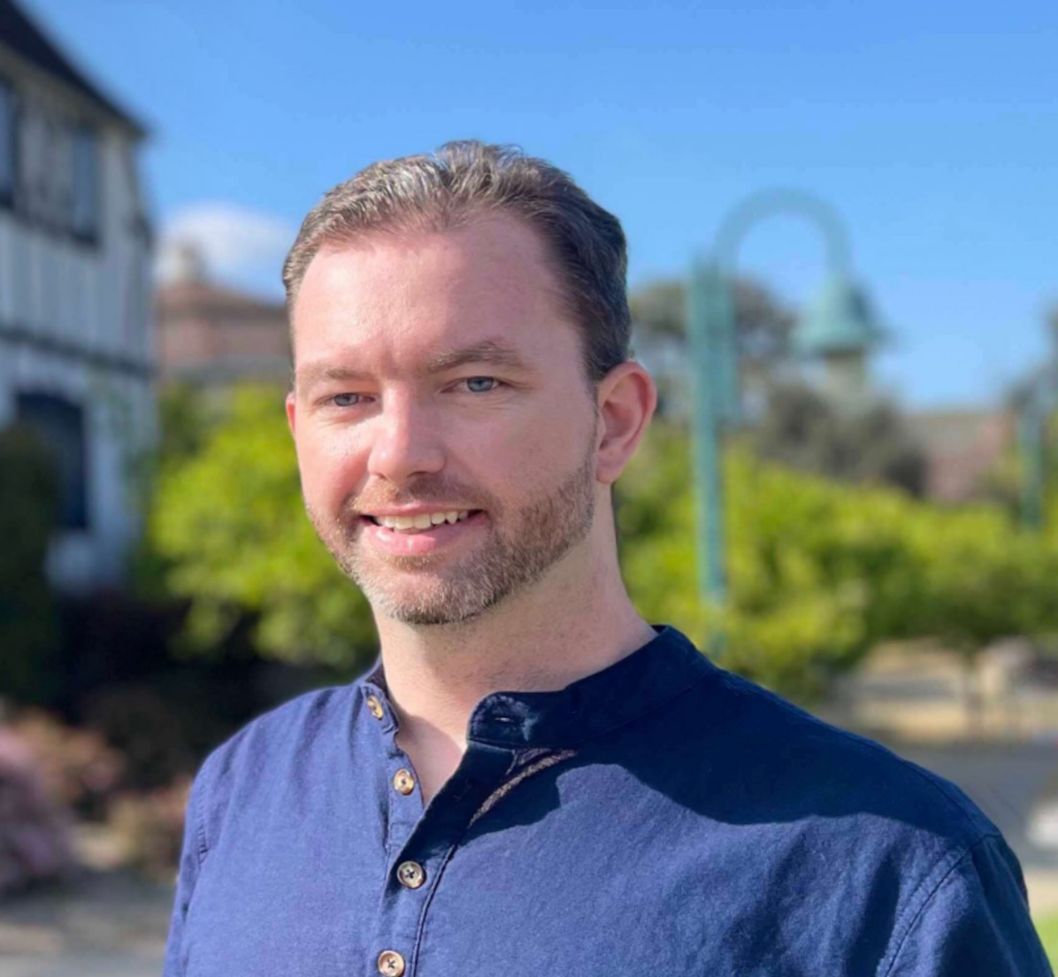 A man smiling outdoors with a blue sky, green trees, and buildings in the background.