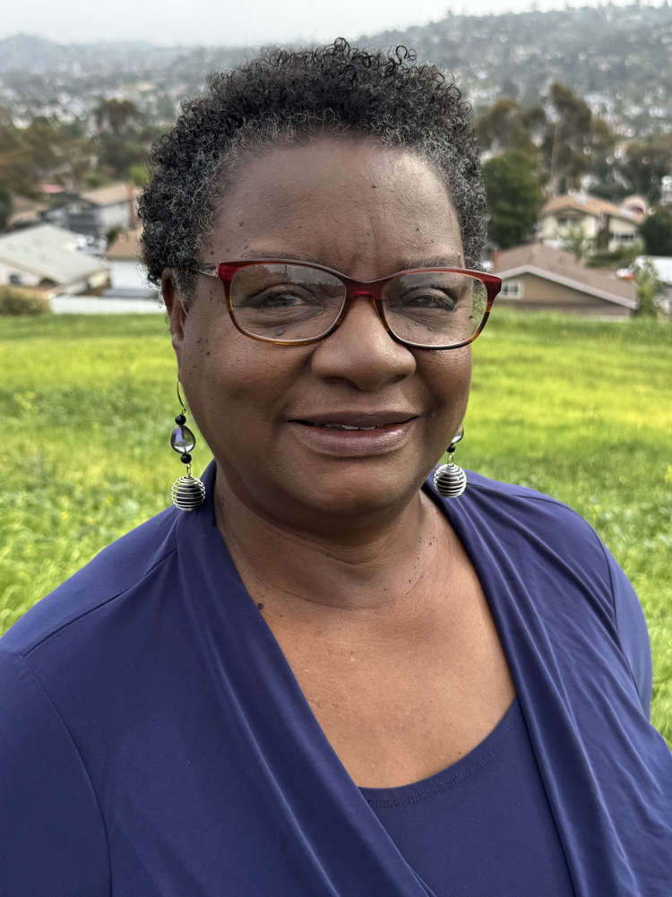 A woman with short, curly hair and glasses outdoors on a grassy hill, with houses and a hilly landscape in the background.
