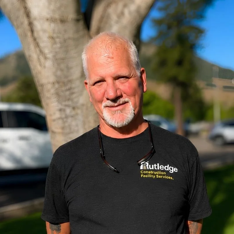 A man with short white hair and a beard, wearing a black T-shirt with the logo 'Rutledge Construction Facility Services,' standing outdoors in front of a large tree with a background of cars and distant trees.