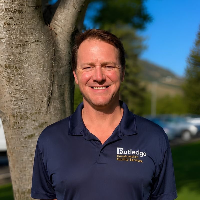 Smiling man standing outdoors near a tree, wearing a navy polo shirt with the logo 'Rutledge Contraction Facility Services'.
