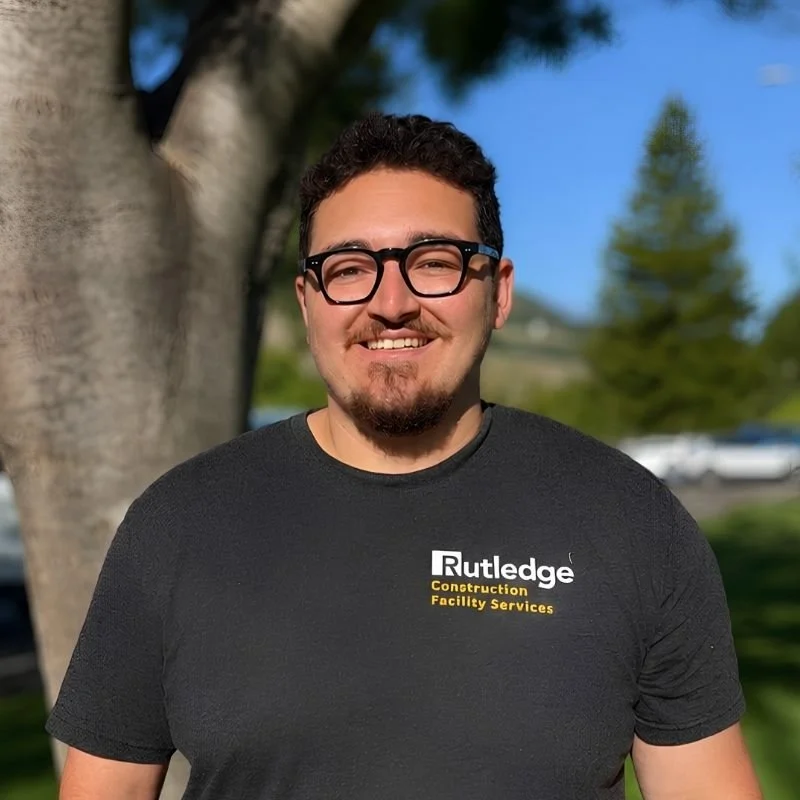 A young man with curly dark hair, glasses, and a goatee smiling outdoors, wearing a black Rutledge Construction Facility Services t-shirt