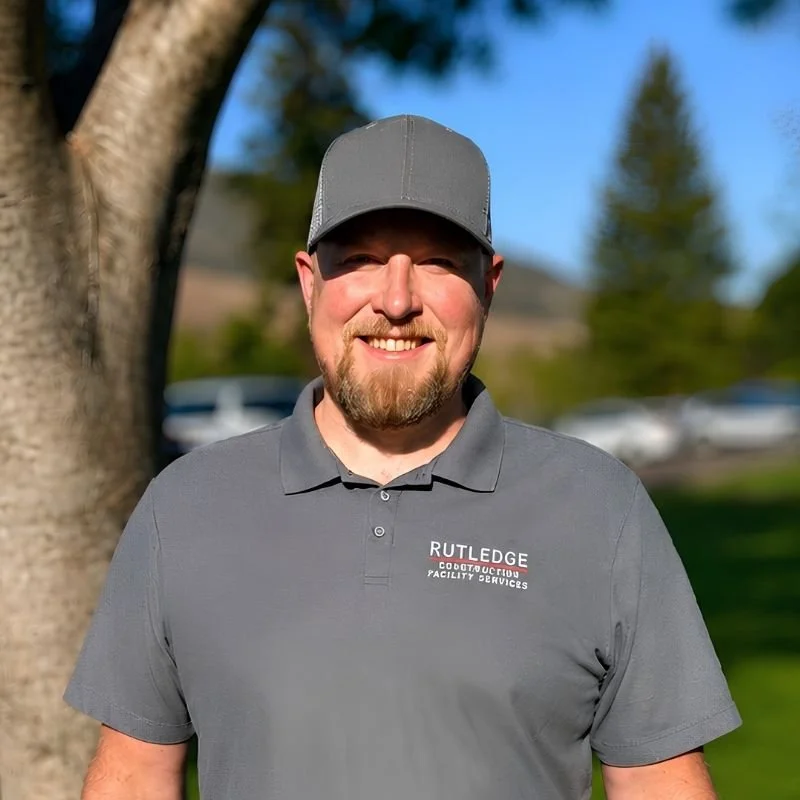 A smiling man wearing a gray polo shirt with 'RUTLEDGE Facility Services' logo and a gray cap, standing outdoors near a tree with blurred cars and trees in the background.