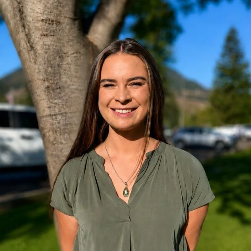 A young woman with long brown hair and a necklace, smiling outdoors in front of a tree, with a blue sky and parked cars in the background.