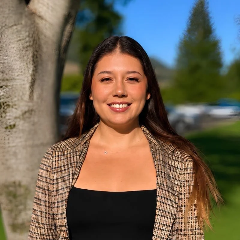 A young woman with long brown hair smiling outdoors near a tree with green foliage and a blue sky.