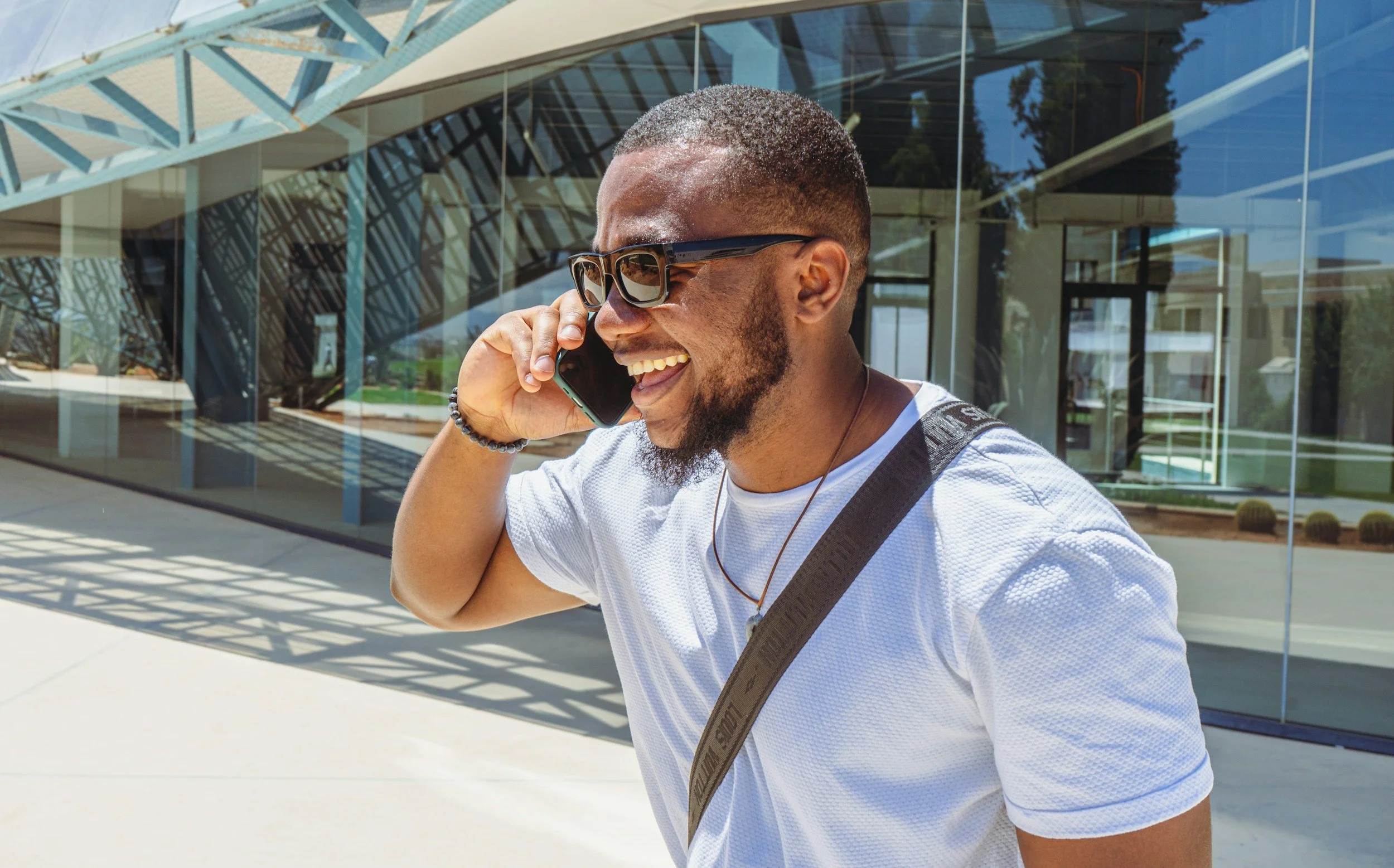 Smiling man with sunglasses talking on a cellphone outside a modern glass building.