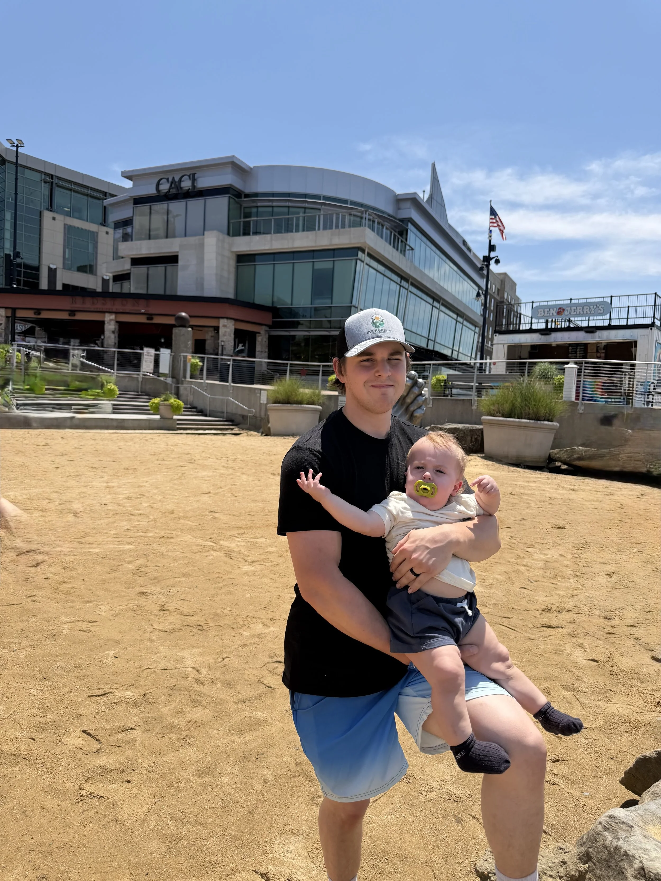A man holding a baby on a sandy beach with buildings and a Ben & Jerry's store in the background, under a clear blue sky.