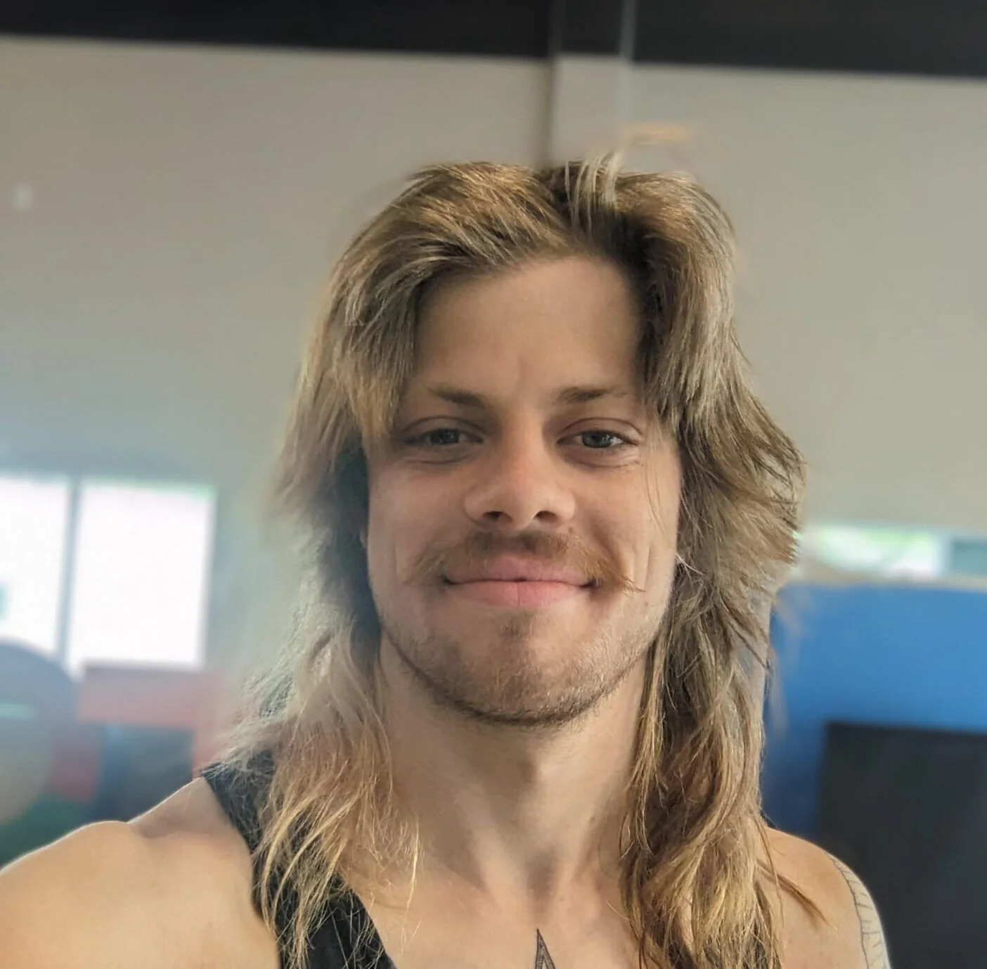 Close-up of a smiling man with long, wavy blonde hair, wearing a sleeveless shirt, in a room with windows in the background.
