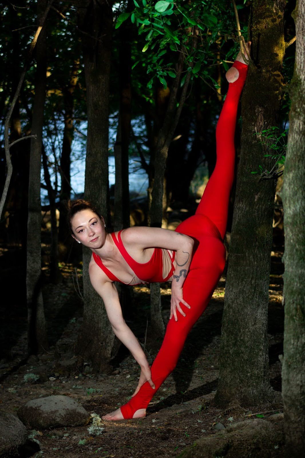 A woman with tattoos wearing a red outfit, doing a yoga pose in a forested area with trees and greenery.