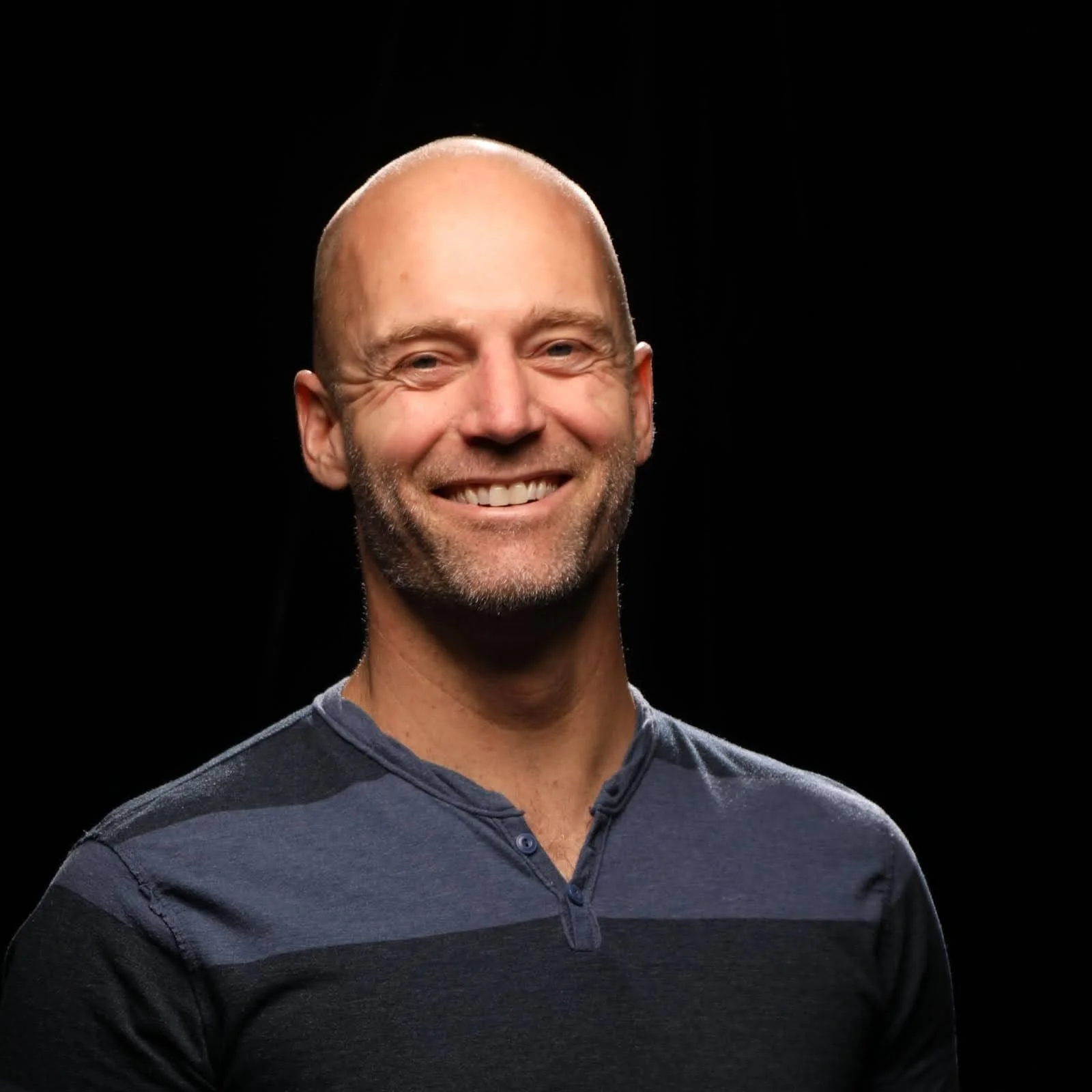 Smiling bald man with a beard wearing a dark blue shirt against a black background.