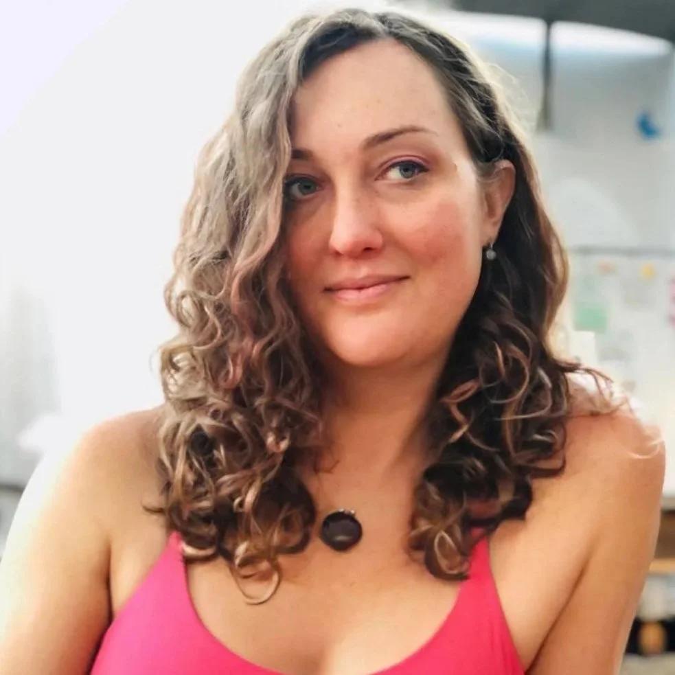 A woman with long, curly brown hair wearing a pink top and earrings, sitting indoors with a whiteboard in the background.