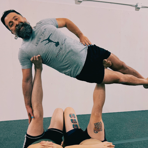 A man with a beard and a gray t-shirt being lifted by a person lying on the ground. The man is smiling and the background is a plain white wall.