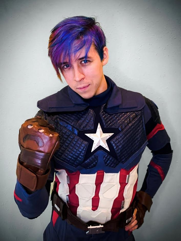 A person dressed as Captain America with purple hair, wearing a costume with a star emblem on the chest and a brown glove, striking a confident pose.