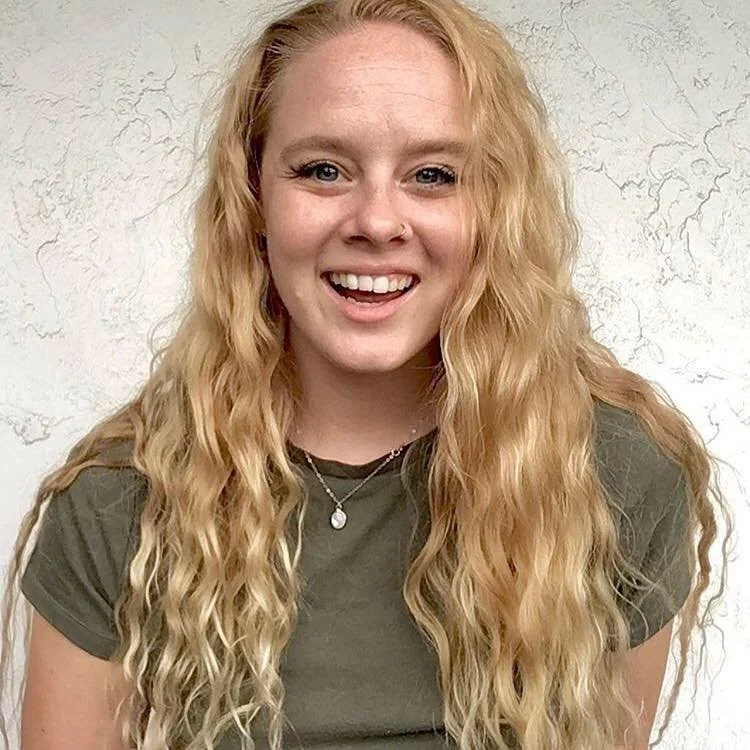 A young woman with long, curly blonde hair smiling in front of a textured white wall.