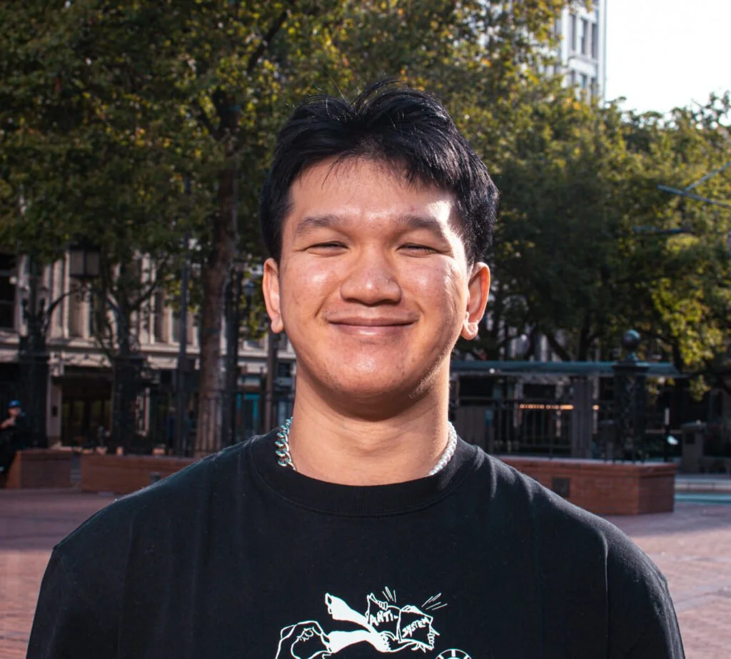 Smiling person with short black hair, wearing a black T-shirt with white graphics, in an outdoor urban setting with trees and buildings in the background.