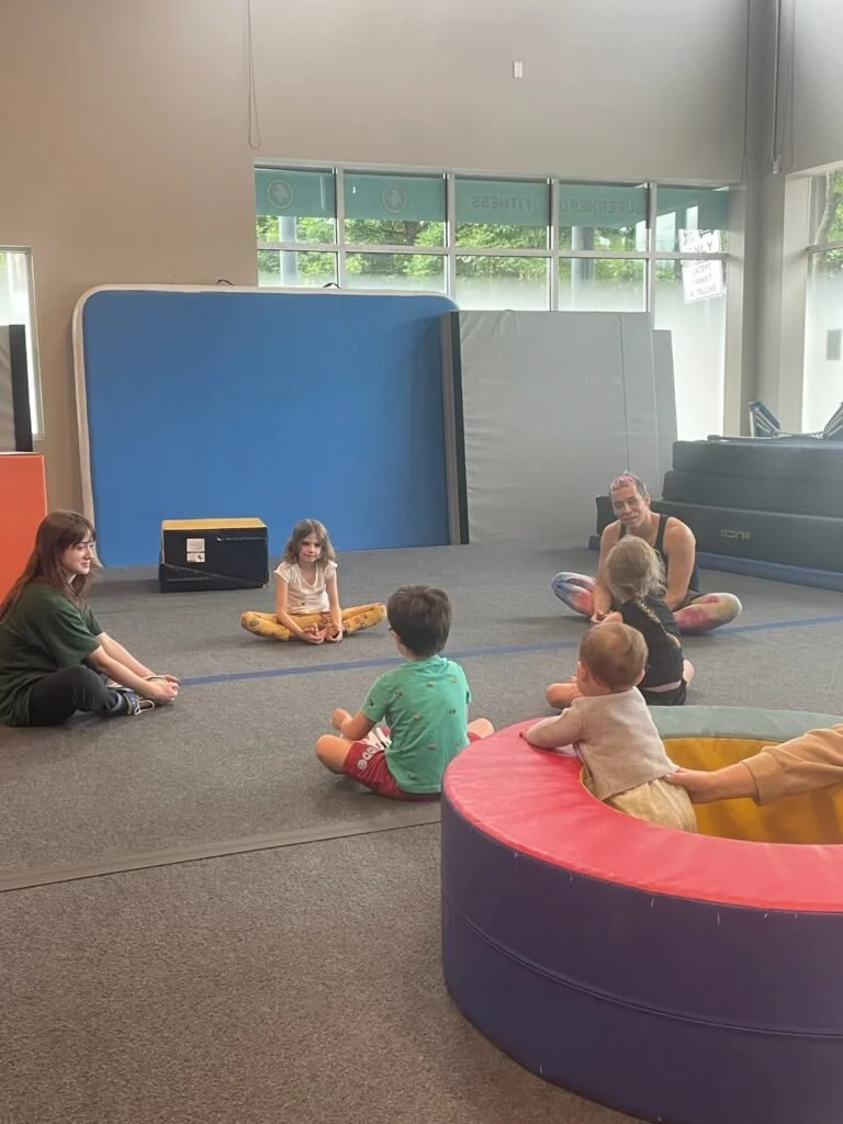 A group of children and a teacher sitting in a circle on a gym floor during a class or activity.