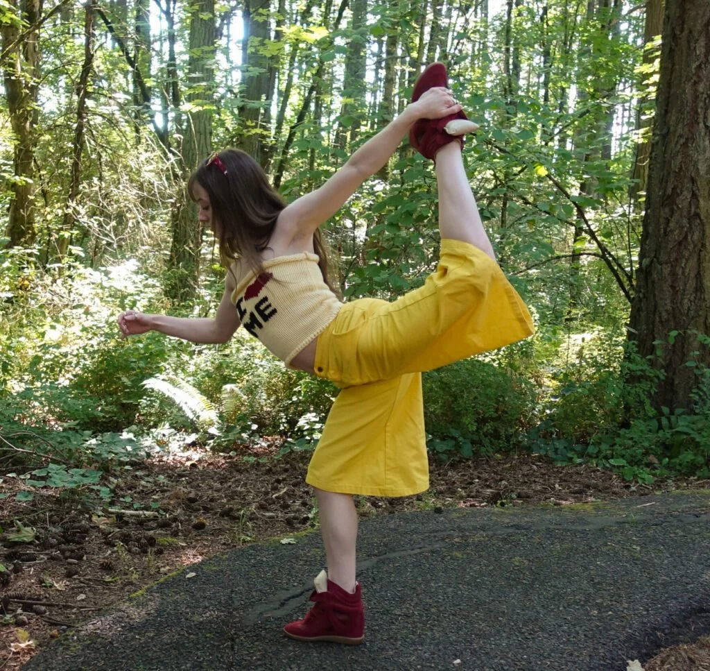 A young woman in a park, standing on one leg, holding her other foot behind her head, and wearing yellow shorts, a sleeveless top, and red boots.