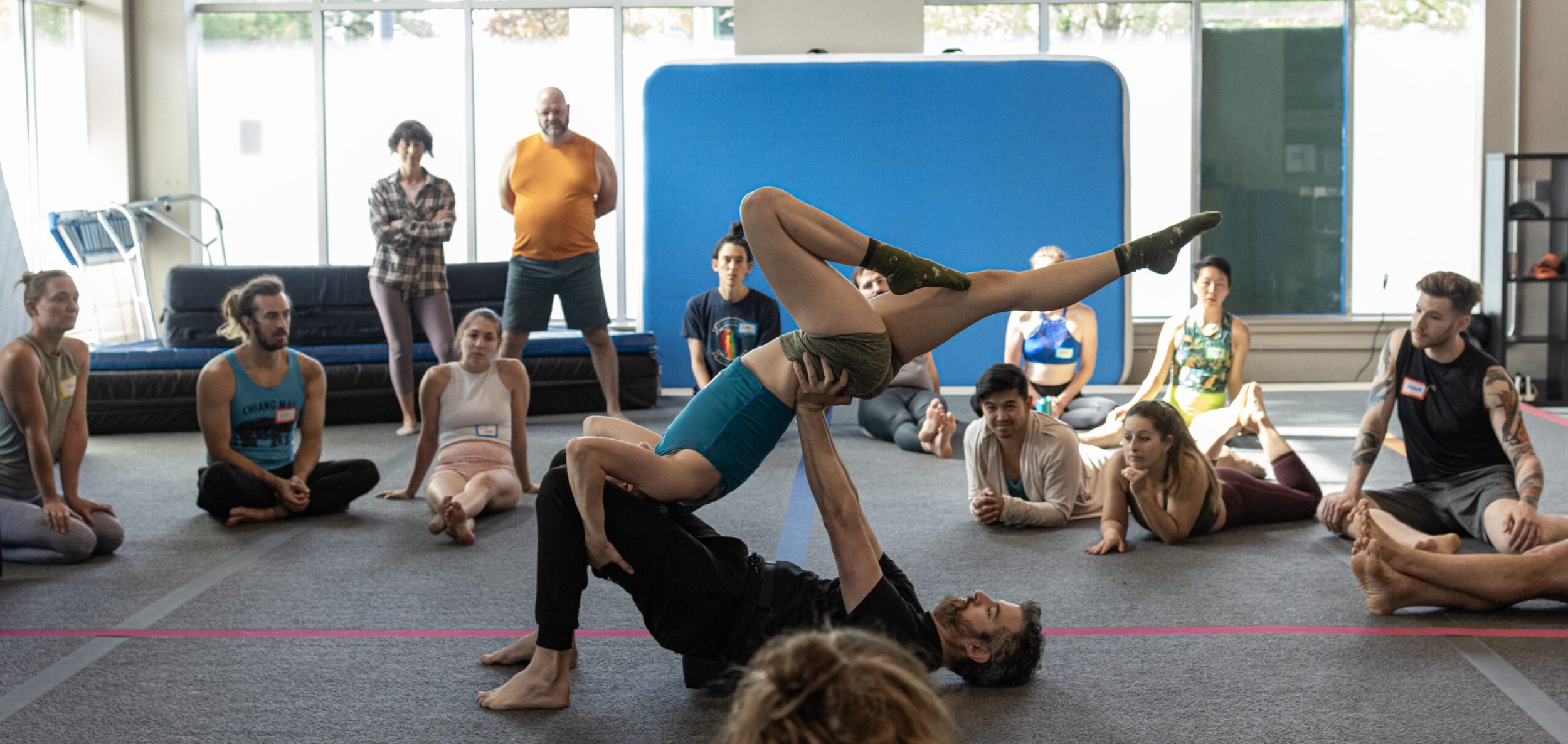 A group of people watching two acrobats performing a balancing act in a spacious room with large windows, some seated on the floor and others standing.