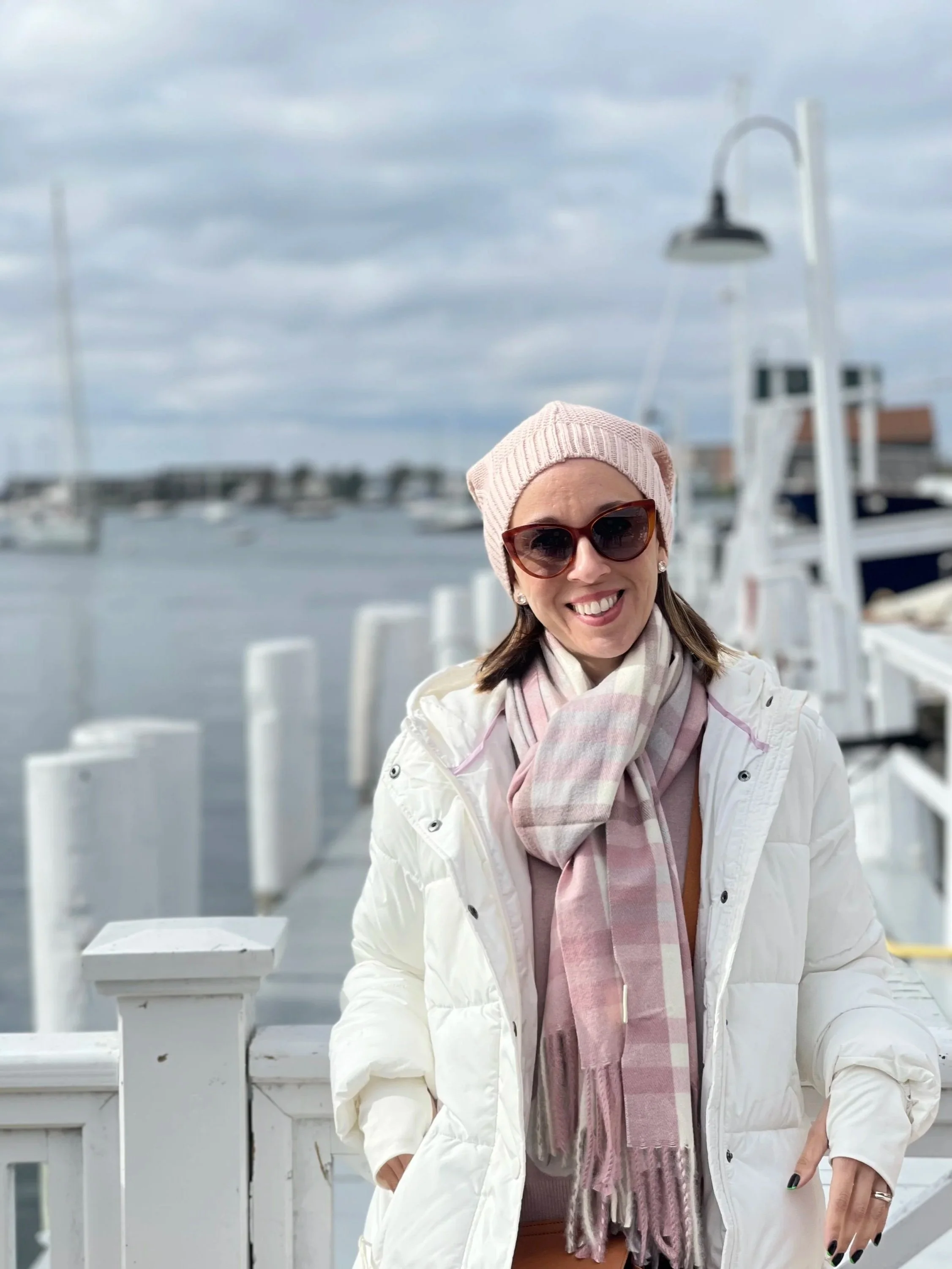 A woman smiling outdoors on a cloudy day, wearing sunglasses, a light pink beanie, a white jacket, and a pink scarf, standing near a marina with boats in the background.