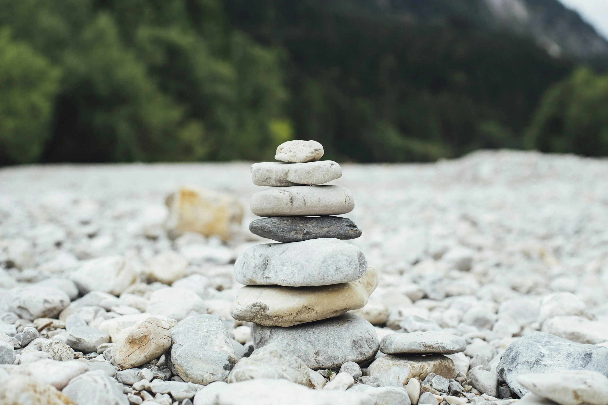 A small stack of six smooth white and gray stones balanced upright on a bed of similar stones, with a blurred green forest in the background.