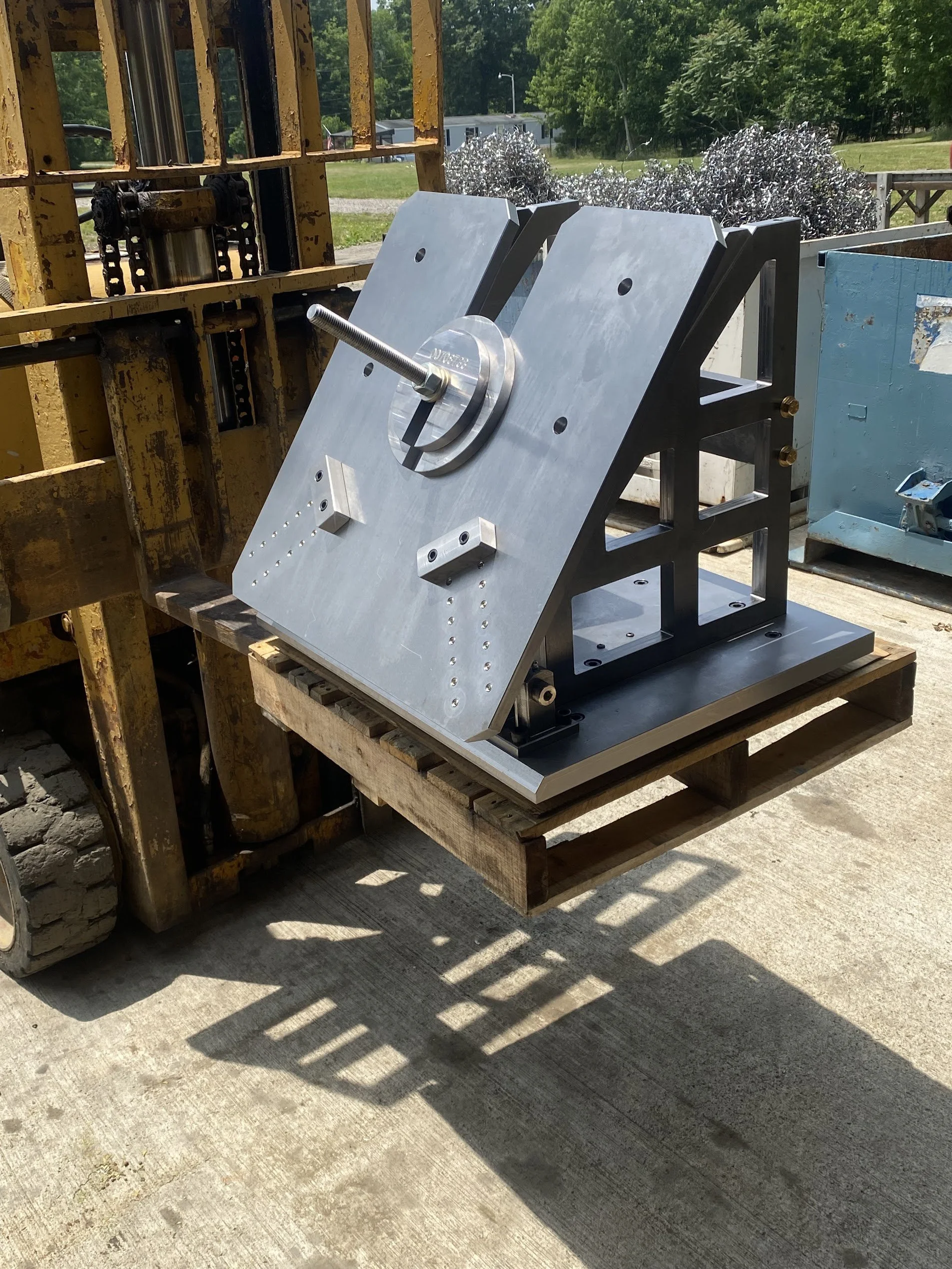 A metal machine part mounted on a black frame outside on a concrete surface, with a yellow rusty pallet and green trees in the background.