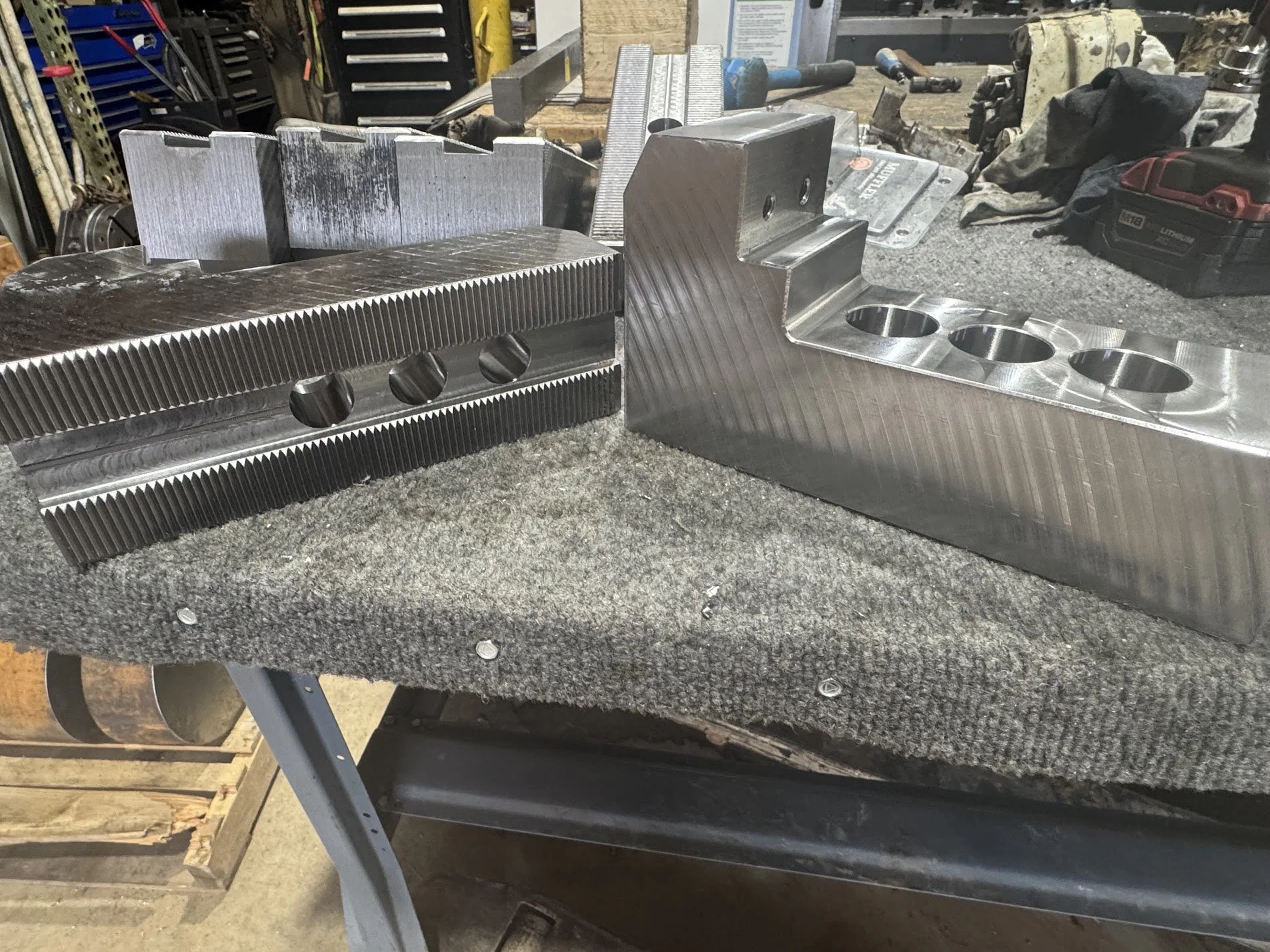 Two large metal CNC machined parts with grooves and holes resting on a workbench in a workshop surrounded by tools and equipment.