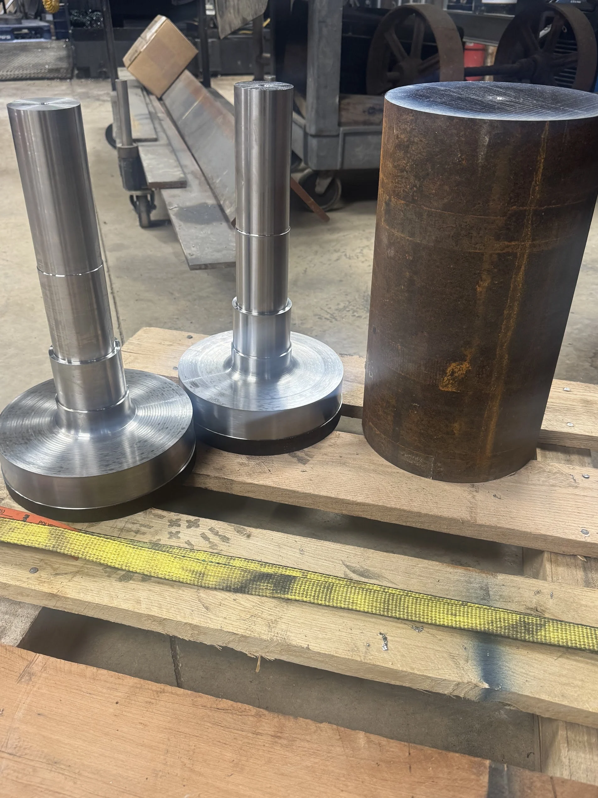 Three cylindrical metal objects with flat bases on a wooden workbench, with a rusty metal cylinder beside them. Tools and equipment are visible in the background.