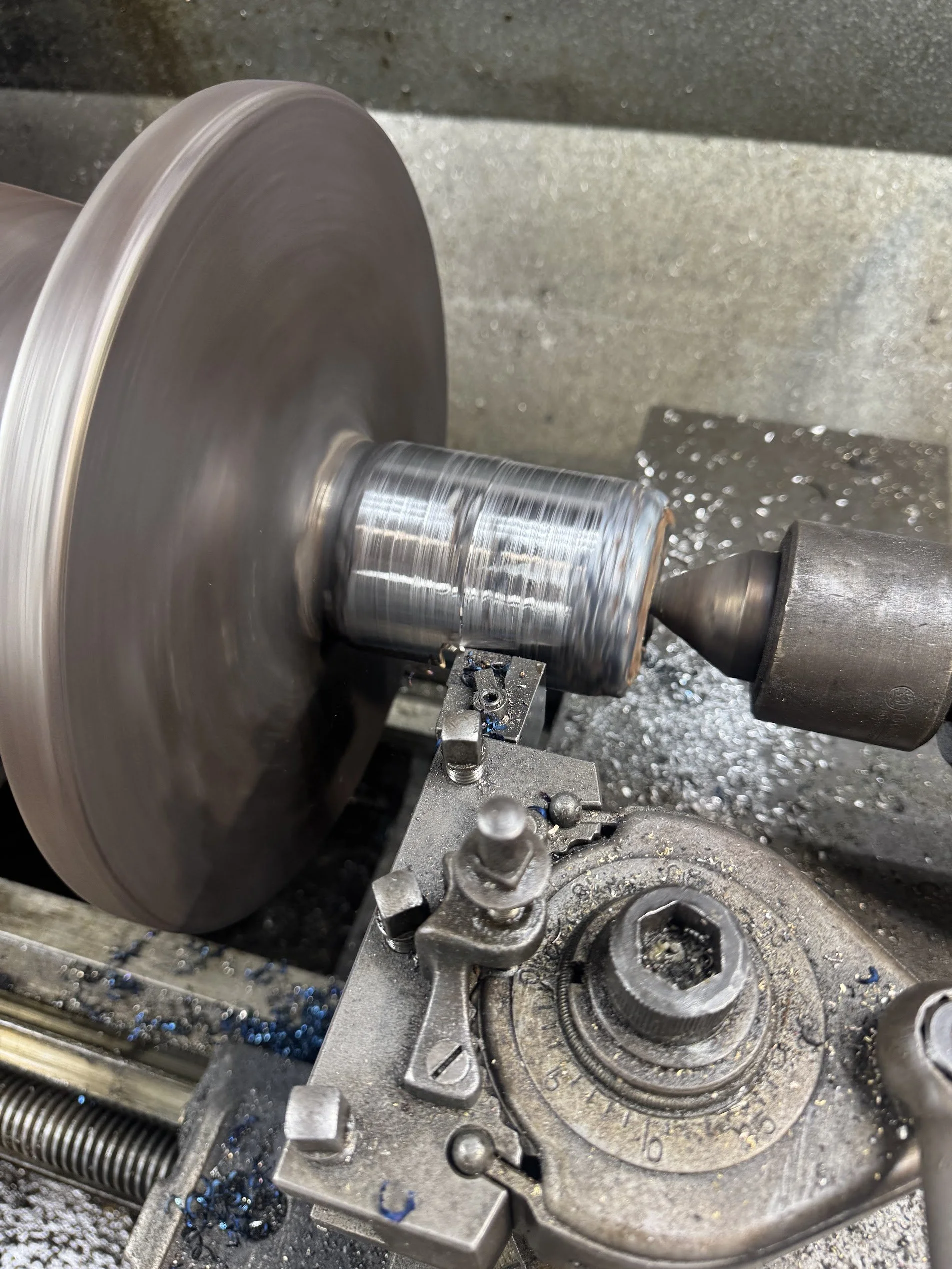 A metal lathe machine shaping a cylindrical metal piece, with metal shavings around and a cutting tool engaged with the metal, creating a shiny surface.