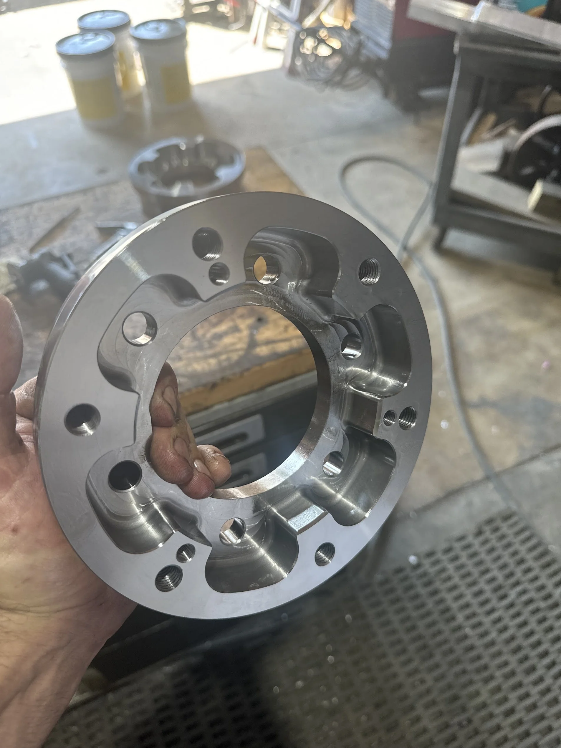 Close-up of a shiny, circular metal mechanical component, possibly a wheel or hub, held in a person's hand in a workshop setting.