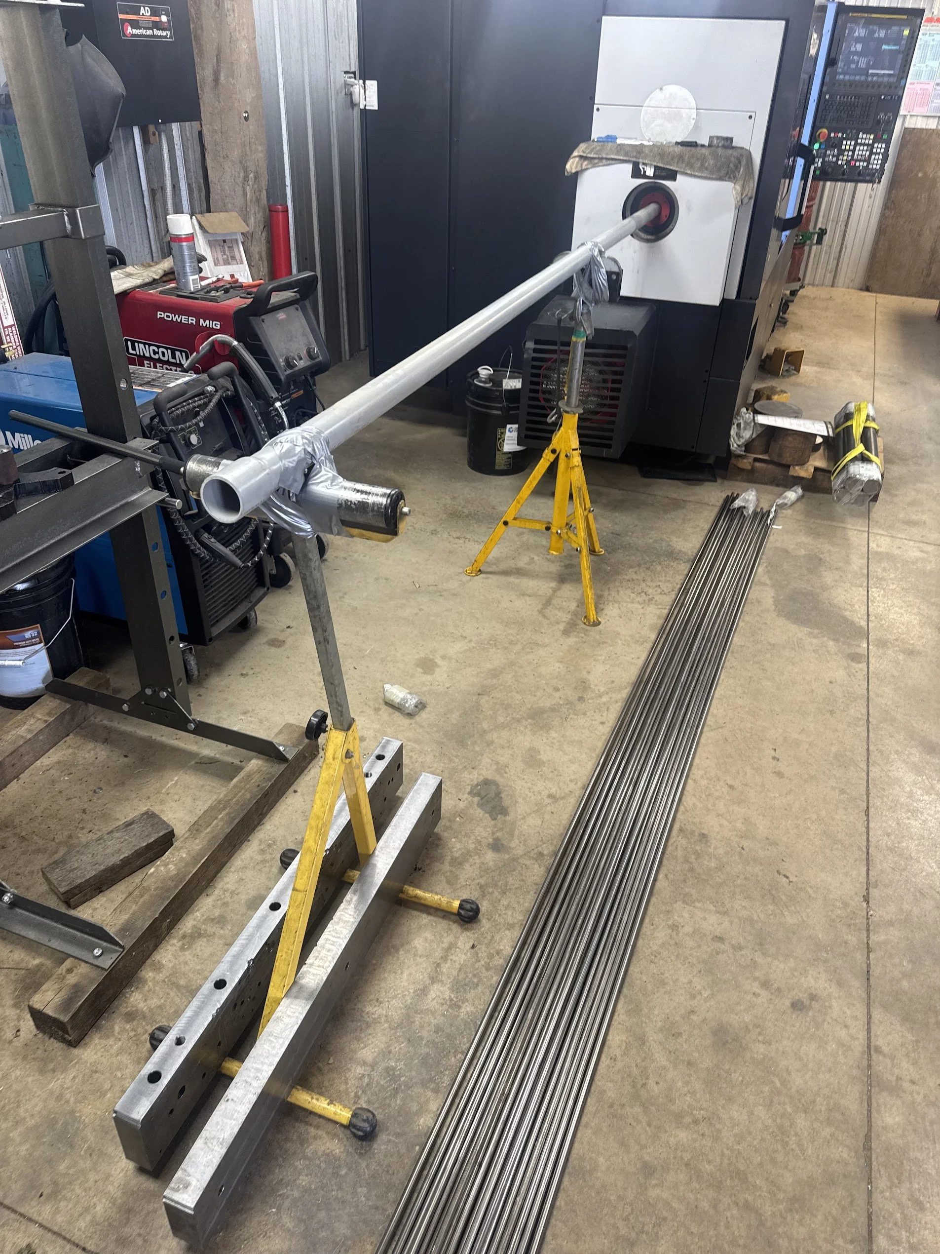 A workshop with a long aluminum pipe placed on a yellow adjustable stand, metal rods lined up on the floor, and welding equipment nearby. The background shows a metal cabinet and a control panel.