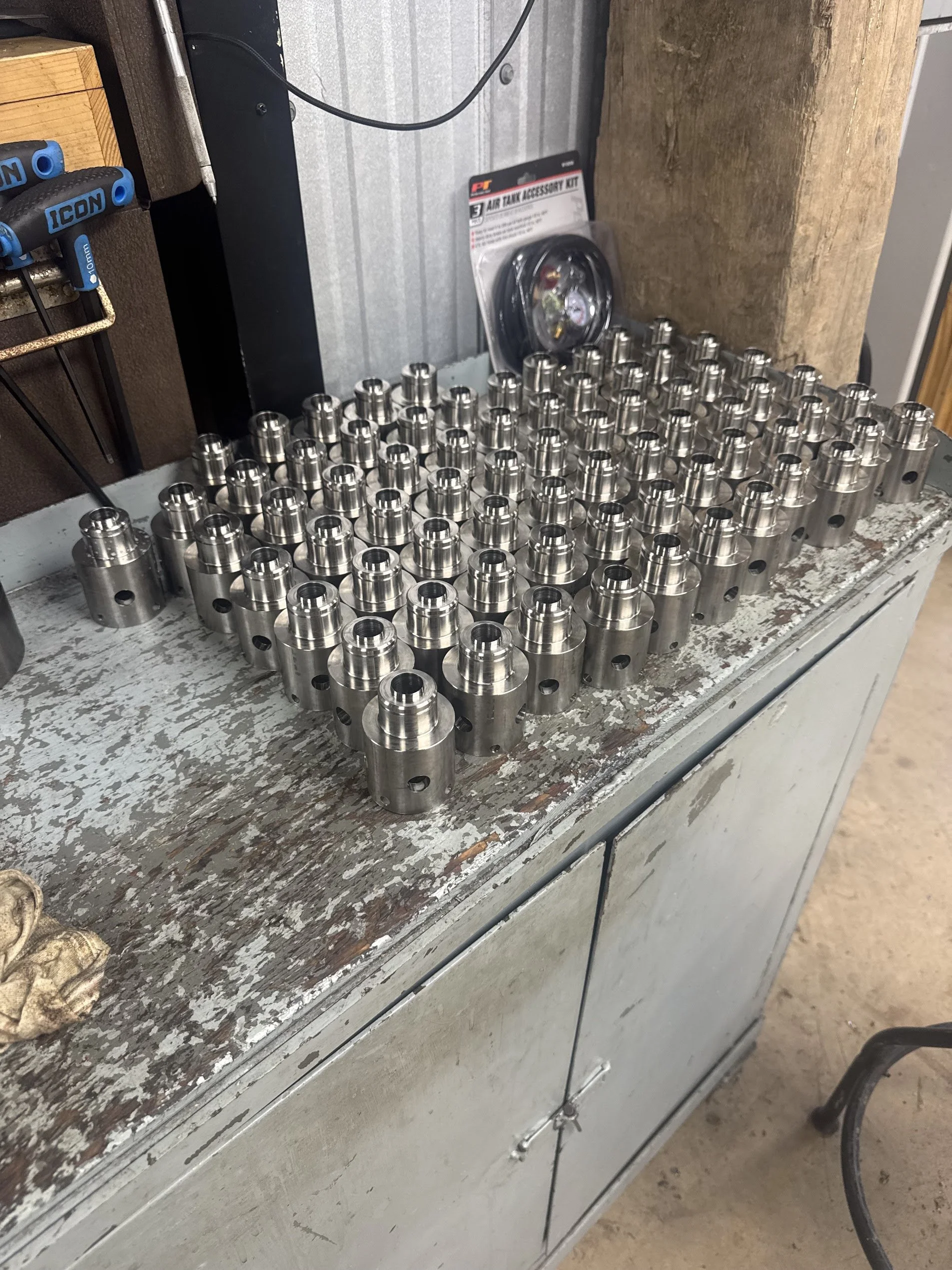 A workbench with a grid of small, metallic cylindrical parts with holes and grooves. To the left, there are screwdrivers and a wire inside a tool holder. A package labeled 'Air Tank Accessory Kit' is on the workbench near a black metal stand, with a 