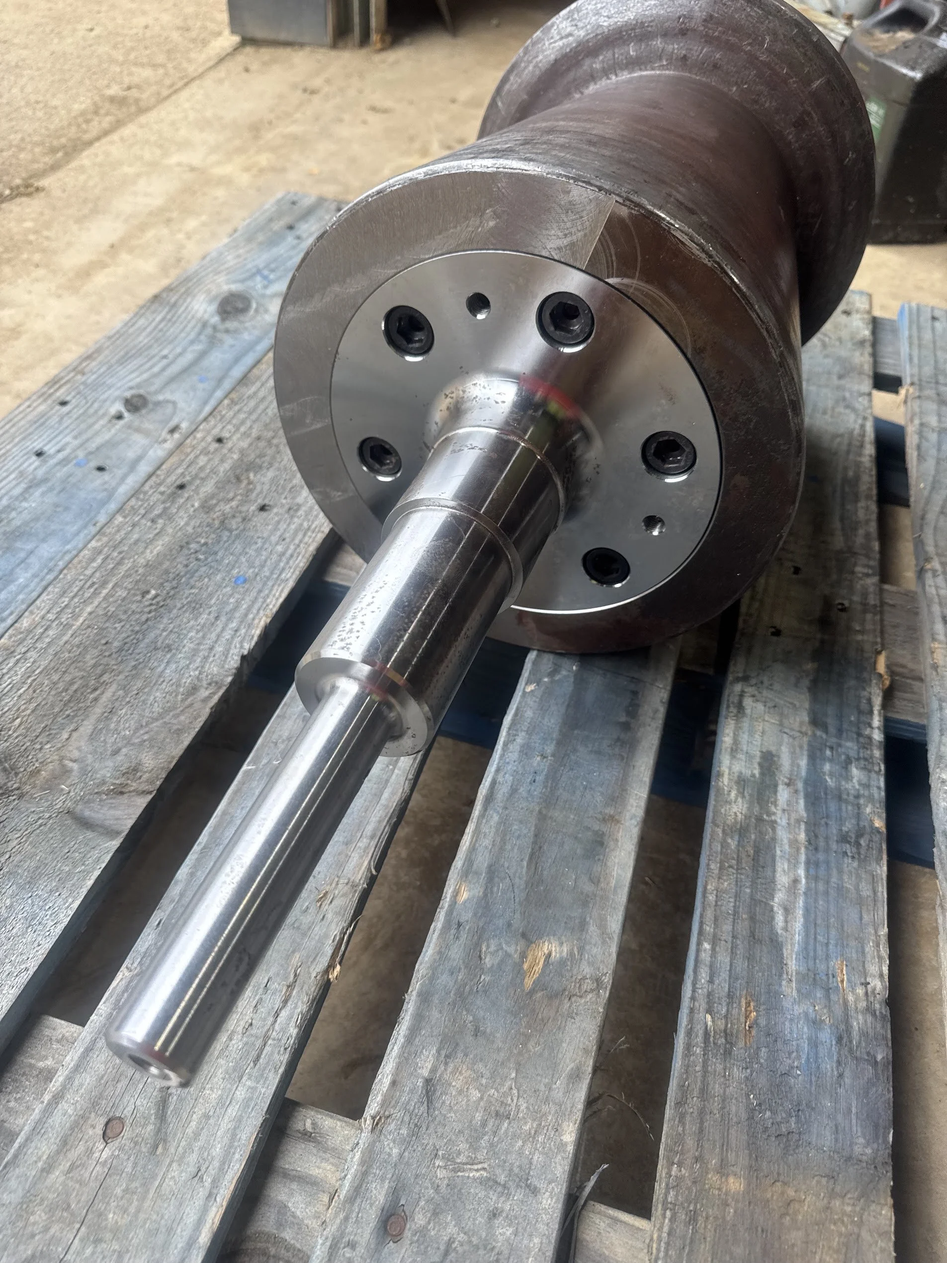 A metal shaft with a flange and bolts resting on a weathered wooden surface, with a workshop or garage setting in the background.