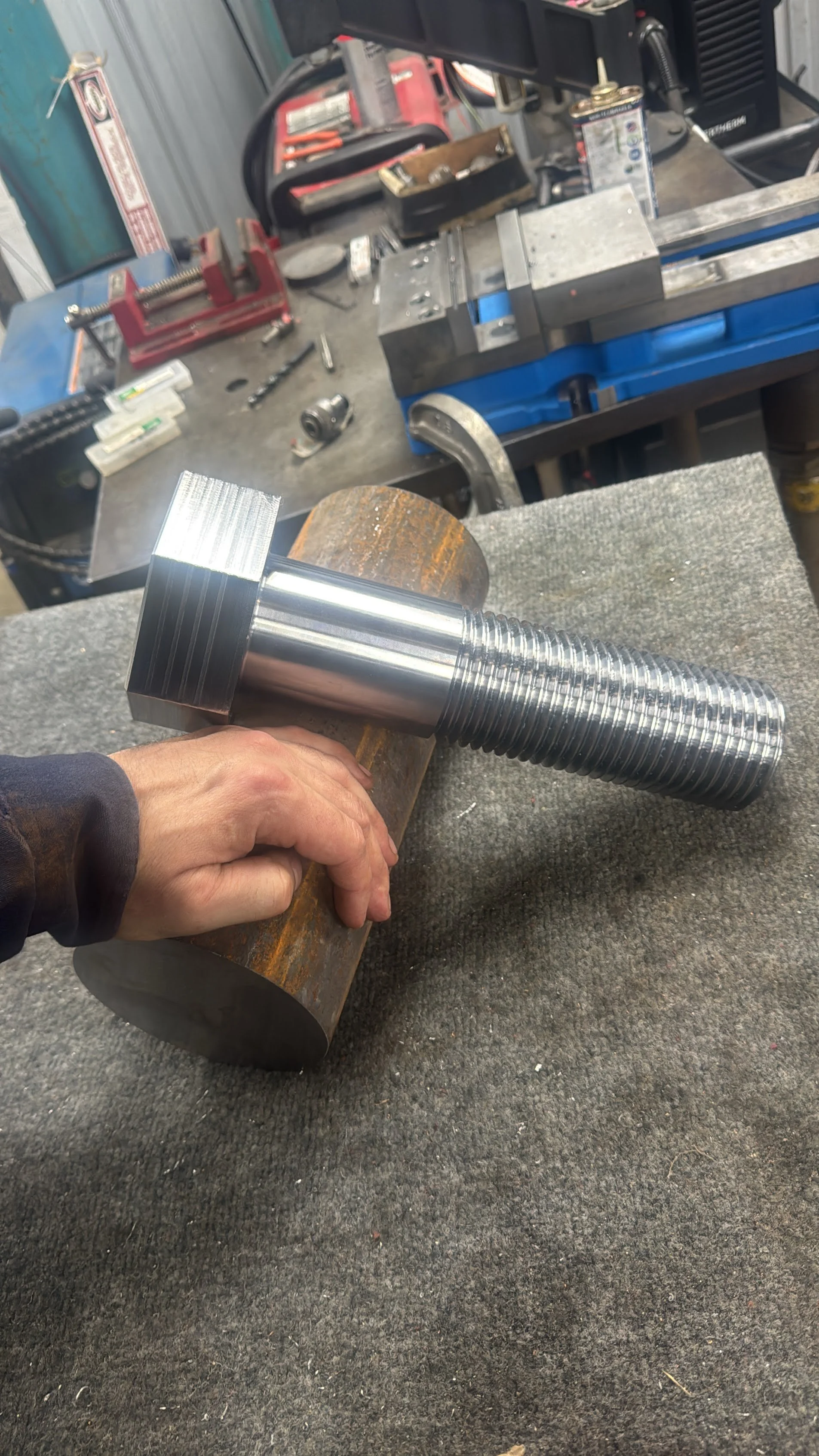 A shiny, new, large metal bolt with a square head and long threaded shaft held in a person's hand against a background of a workbench with tools and equipment in a workshop.