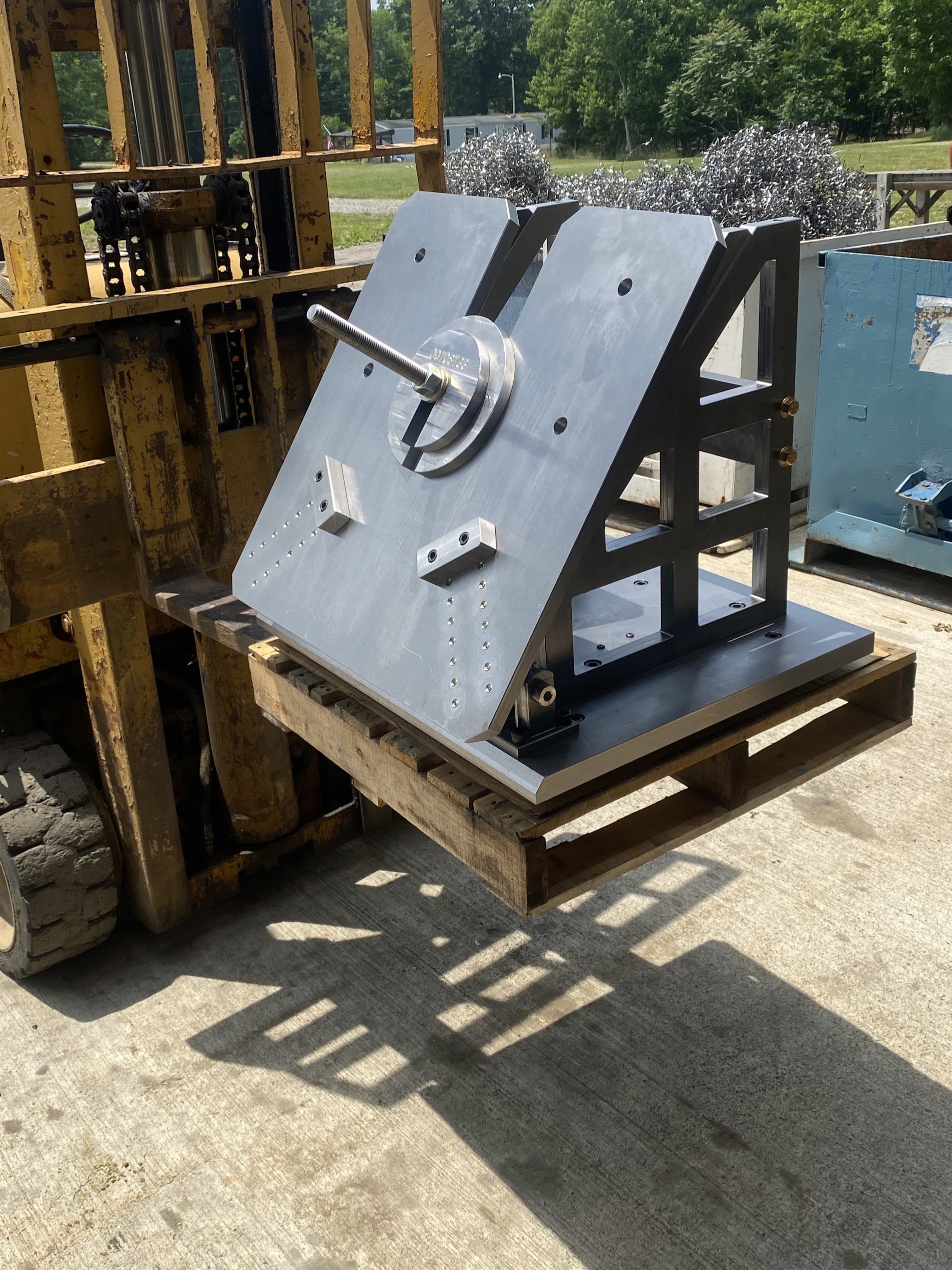 Metal mechanical component on a workbench outdoors, with trees and trailers in the background.