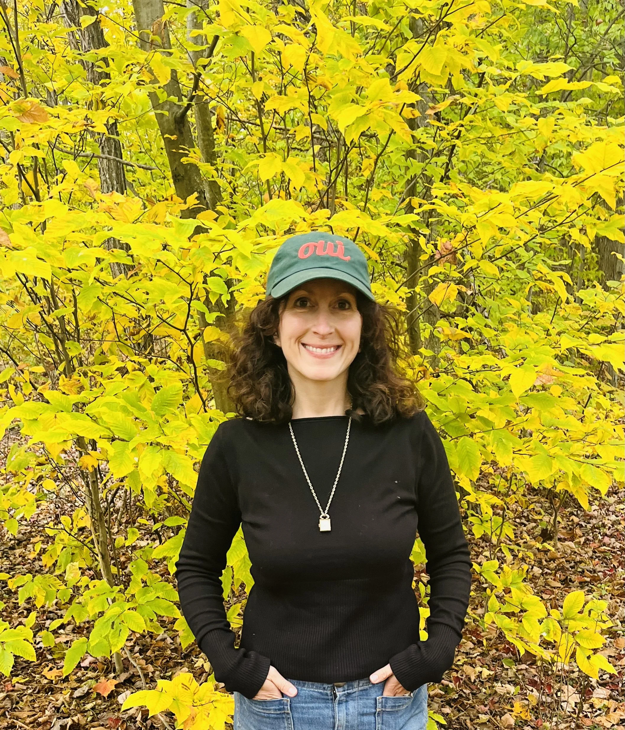 A woman with curly brown hair smiling, wearing a green baseball cap with red writing, a black long-sleeve shirt, and blue jeans, standing outdoors in front of yellow autumn leaves on trees.