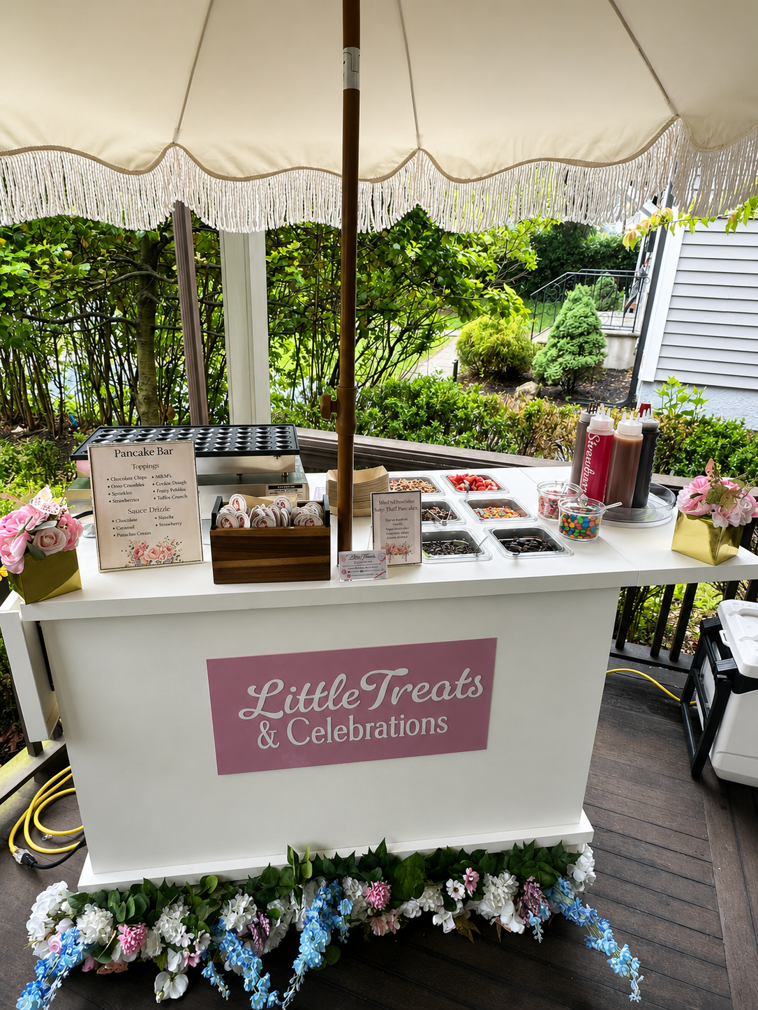 An outdoor pancake and treat stand with a large white umbrella, decorated with flowers at the base, featuring a pink sign that says 'Little Treats & Celebrations.' The stand has various toppings, sprinkles, and syrups, along with a small pancake grid