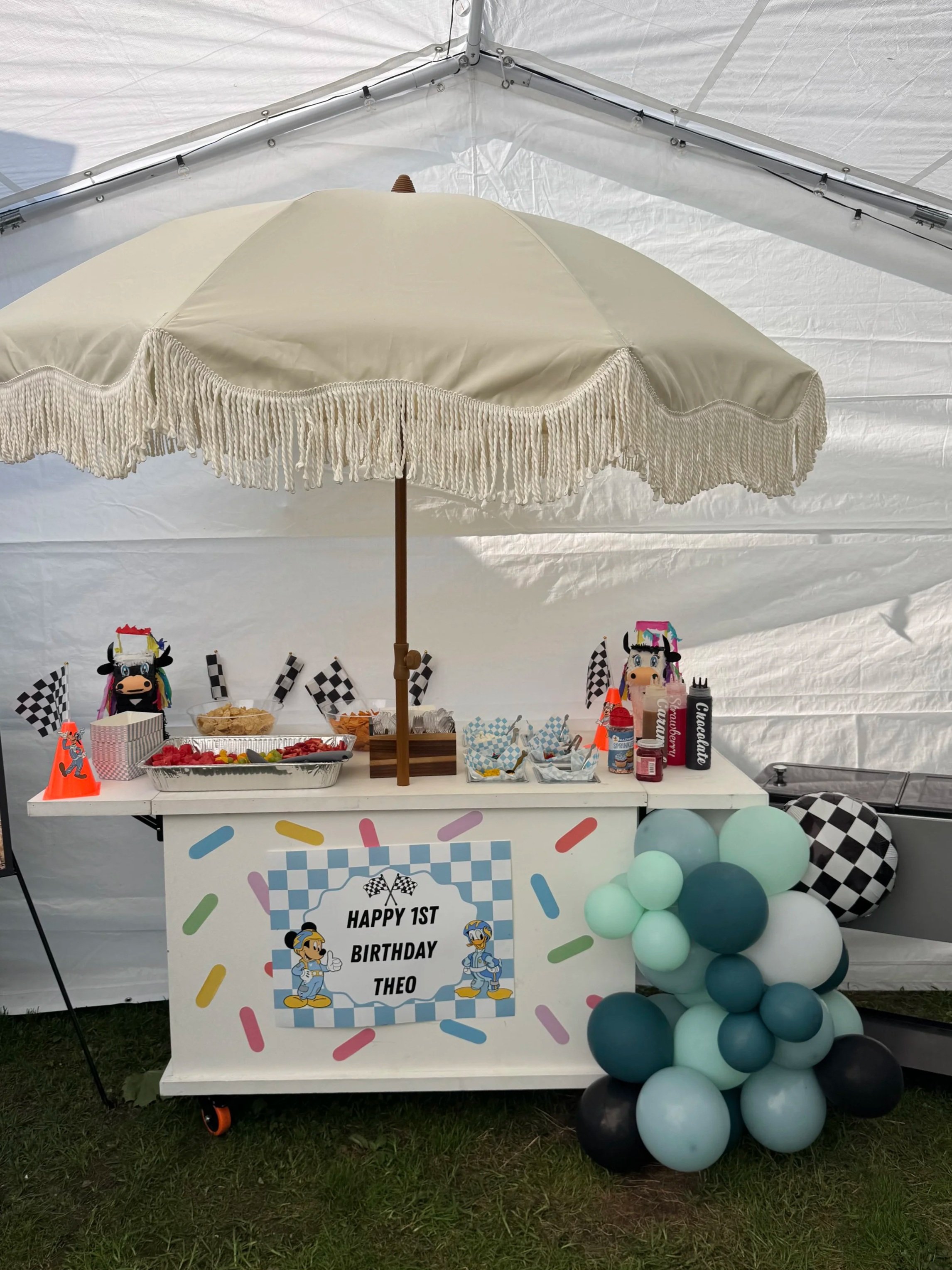 A decorated table for a first birthday party with a sign that reads "Happy 1st Birthday Theo." The table is under a white canopy with a cream-colored umbrella providing shade. Decorations include checkered flags, balloons in shades of blue, teal, and
