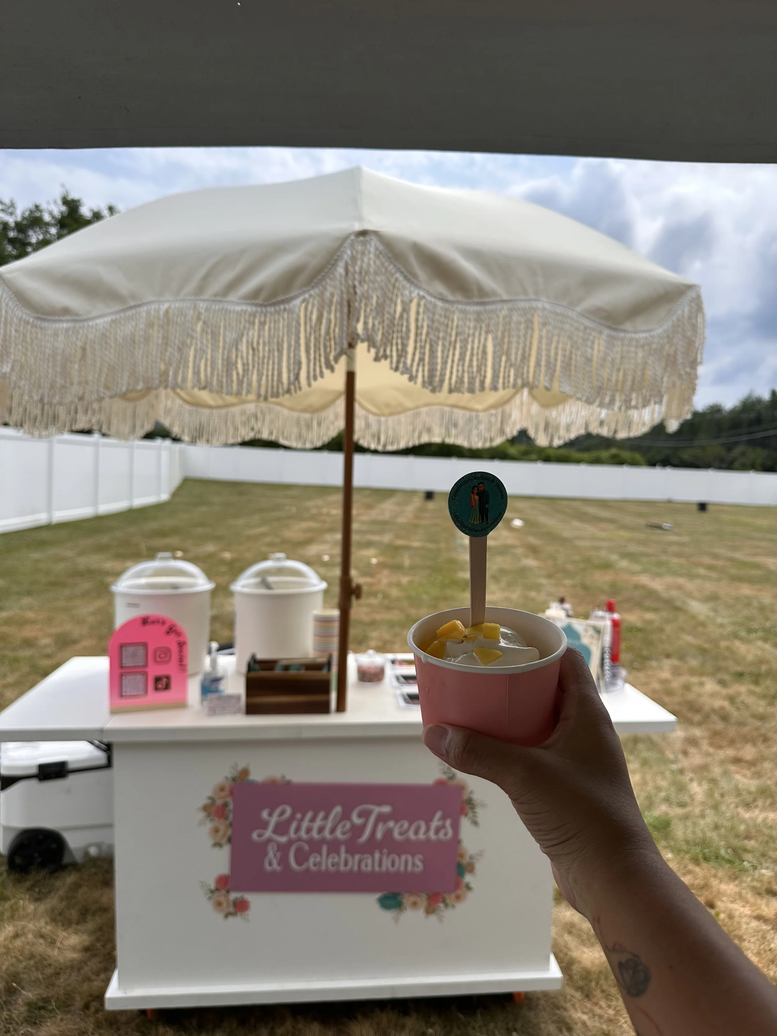 Person holding a cup of frozen treat under a white fringe parasol at an outdoor event with a sign for Little Treats & Celebrations in the background.