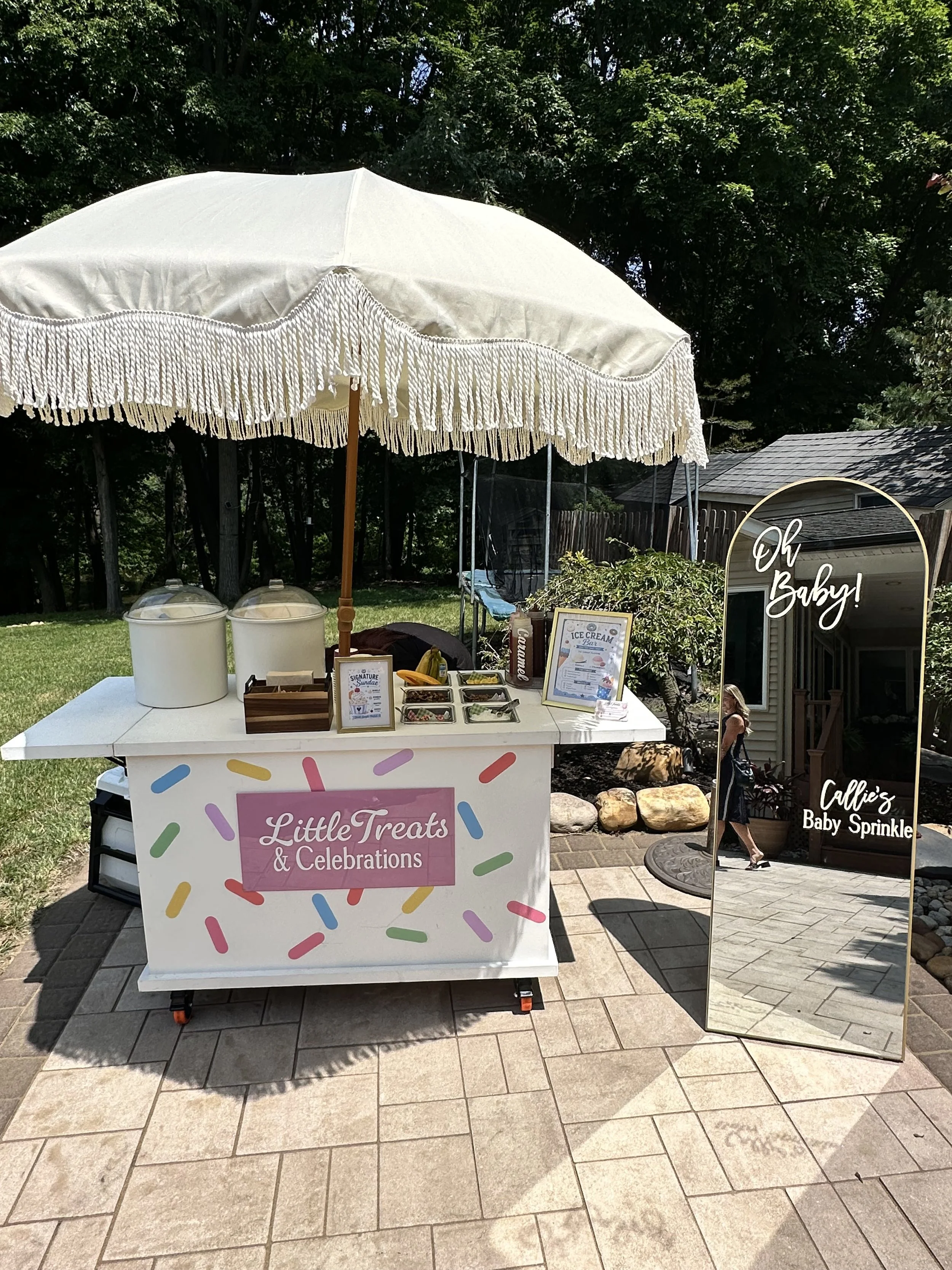 A mobile dessert stand with a pink sign that reads 'Little Treats & Celebrations', decorated with colorful sprinkles, and selling ice cream treats. There is a large white parasol with fringe over the stand. To the right, a woman is walking past a sig