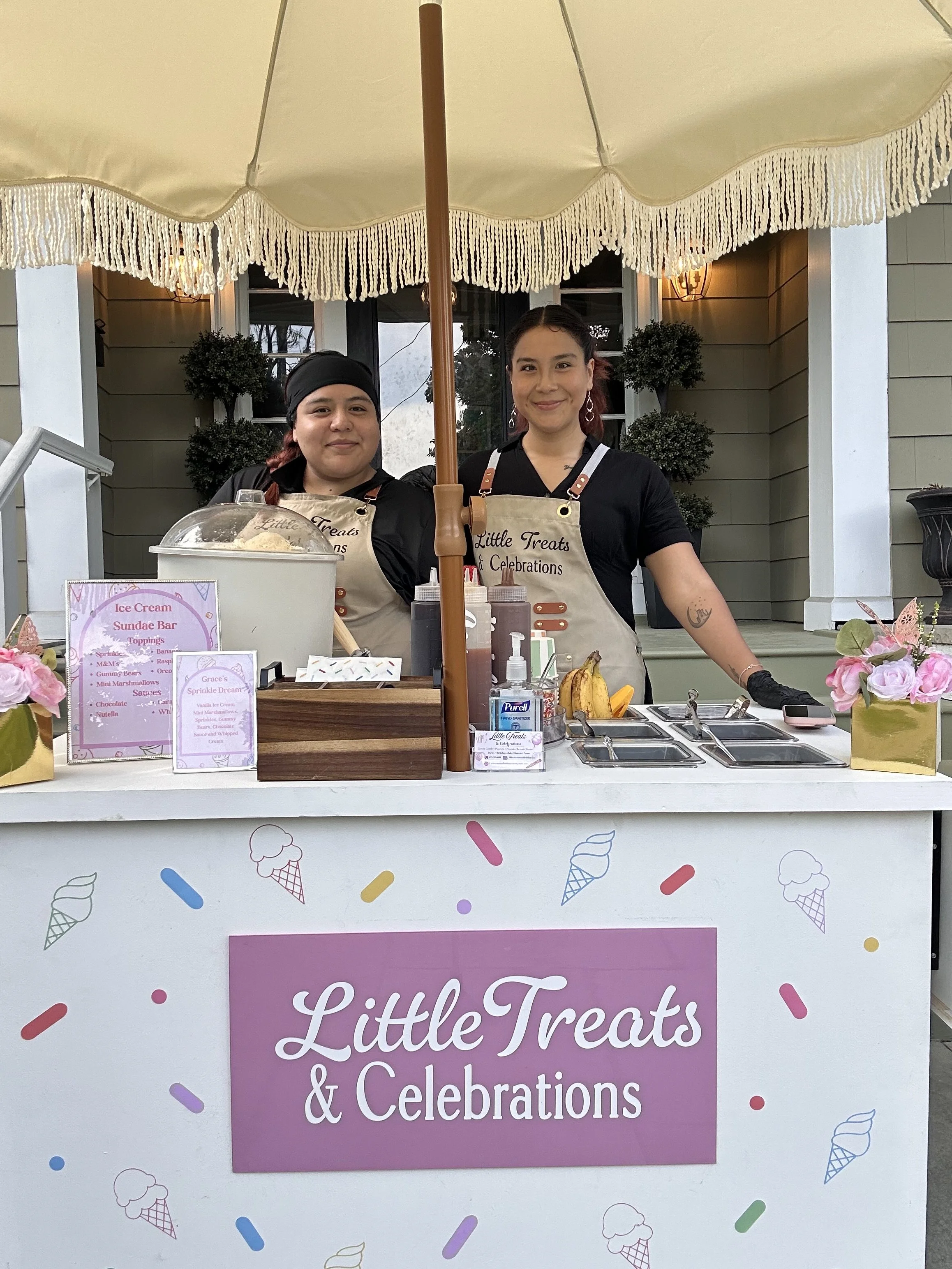 Two women standing behind a dessert stand under a large cream-colored umbrella. The stand has a pink sign that reads 'Little Treats & Celebrations' with colorful ice cream and sprinkles decorations. One woman wears a black headscarf, and the other has red hair. Both are wearing beige aprons.