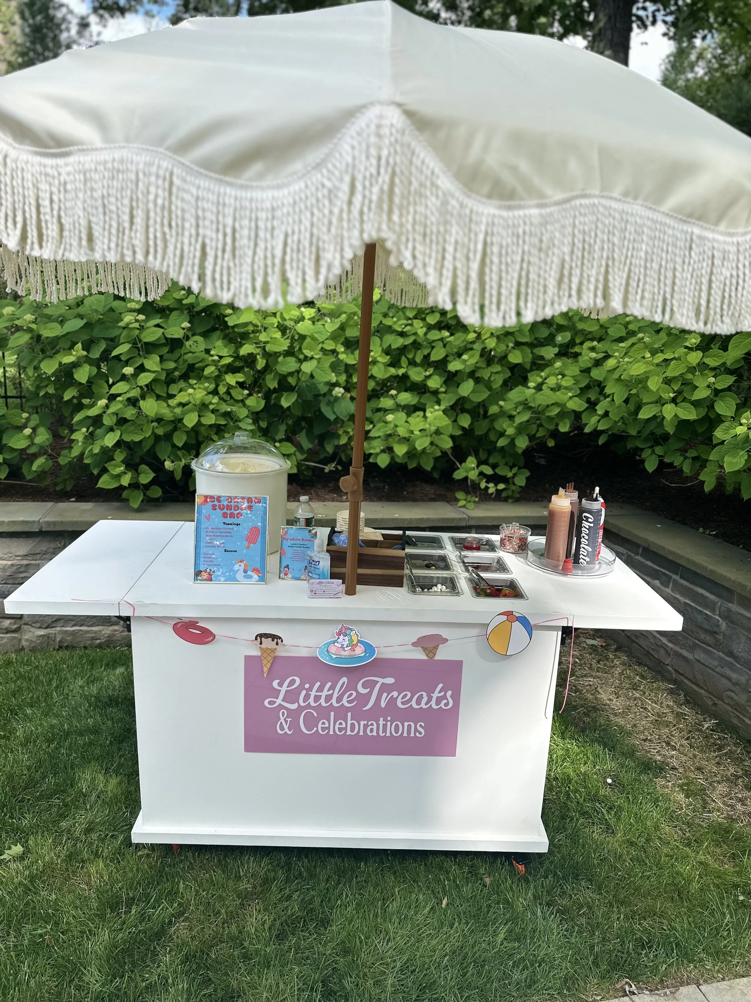 Ice cream stand with a pink sign that reads 'Little Treats & Celebrations,' decorated with images of ice cream cones, a unicorn, and a beach ball, set under a large white umbrella on a grassy area.