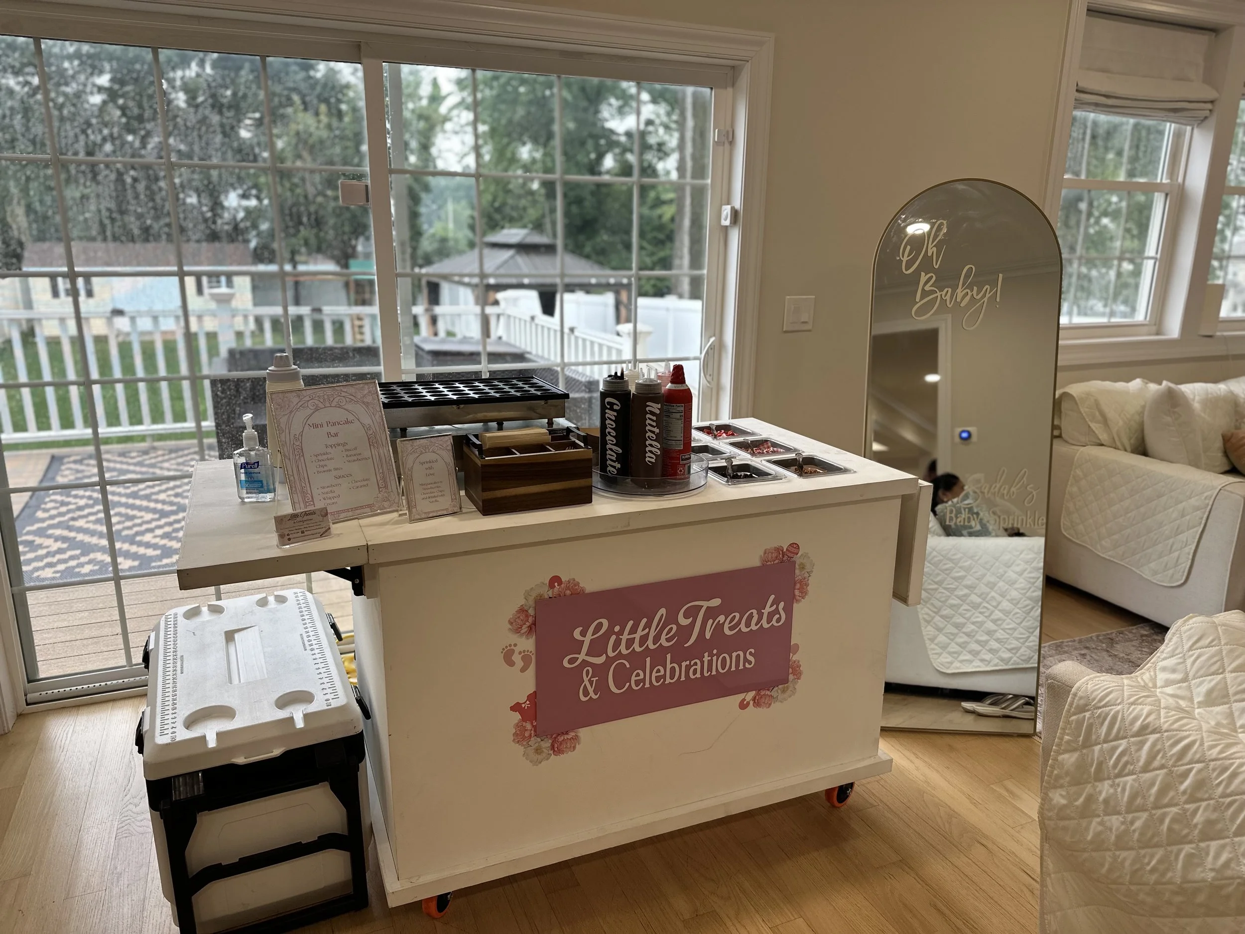 A white cart with a sign reading 'Little Treats & Celebrations,' topped with a pink floral design, and surrounded by a white baby thermometer, chocolate toppings, and a sign menu for mini pancakes. The cart is near a large window showing an outdoor p